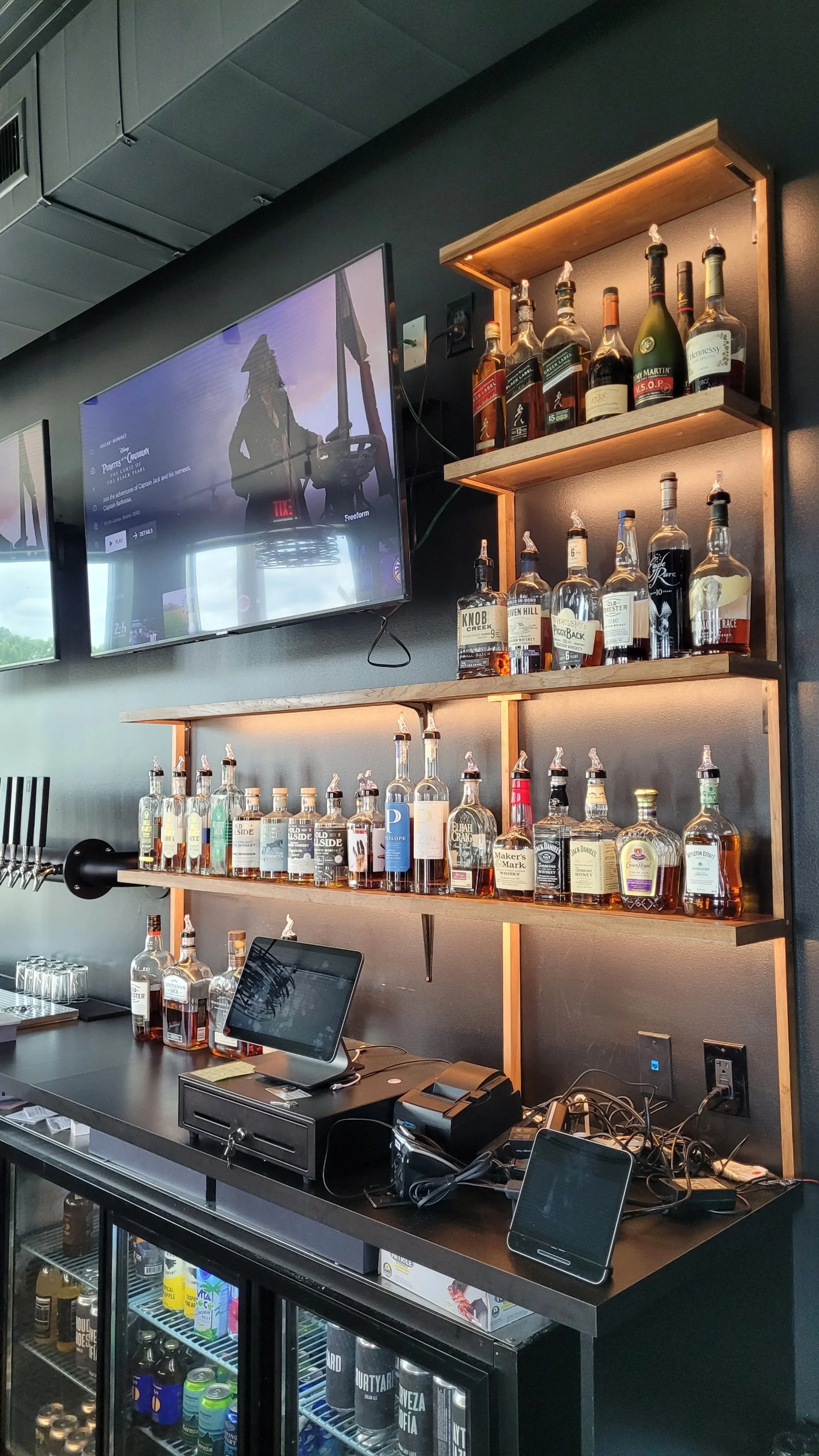 Bar area with shelves of liquor bottles, a TV screen, and electronic equipment on the counter.