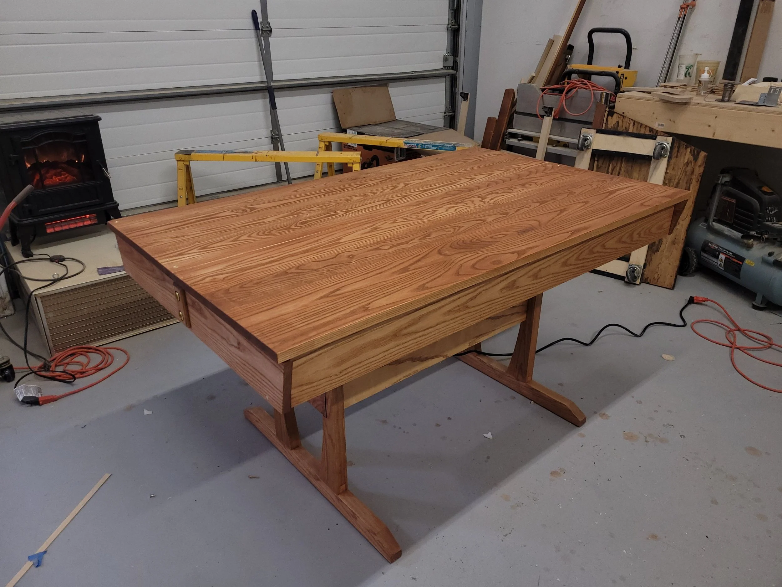 A wooden table in a workshop with various tools and equipment around it, including a heater, power cords, and a workbench.
