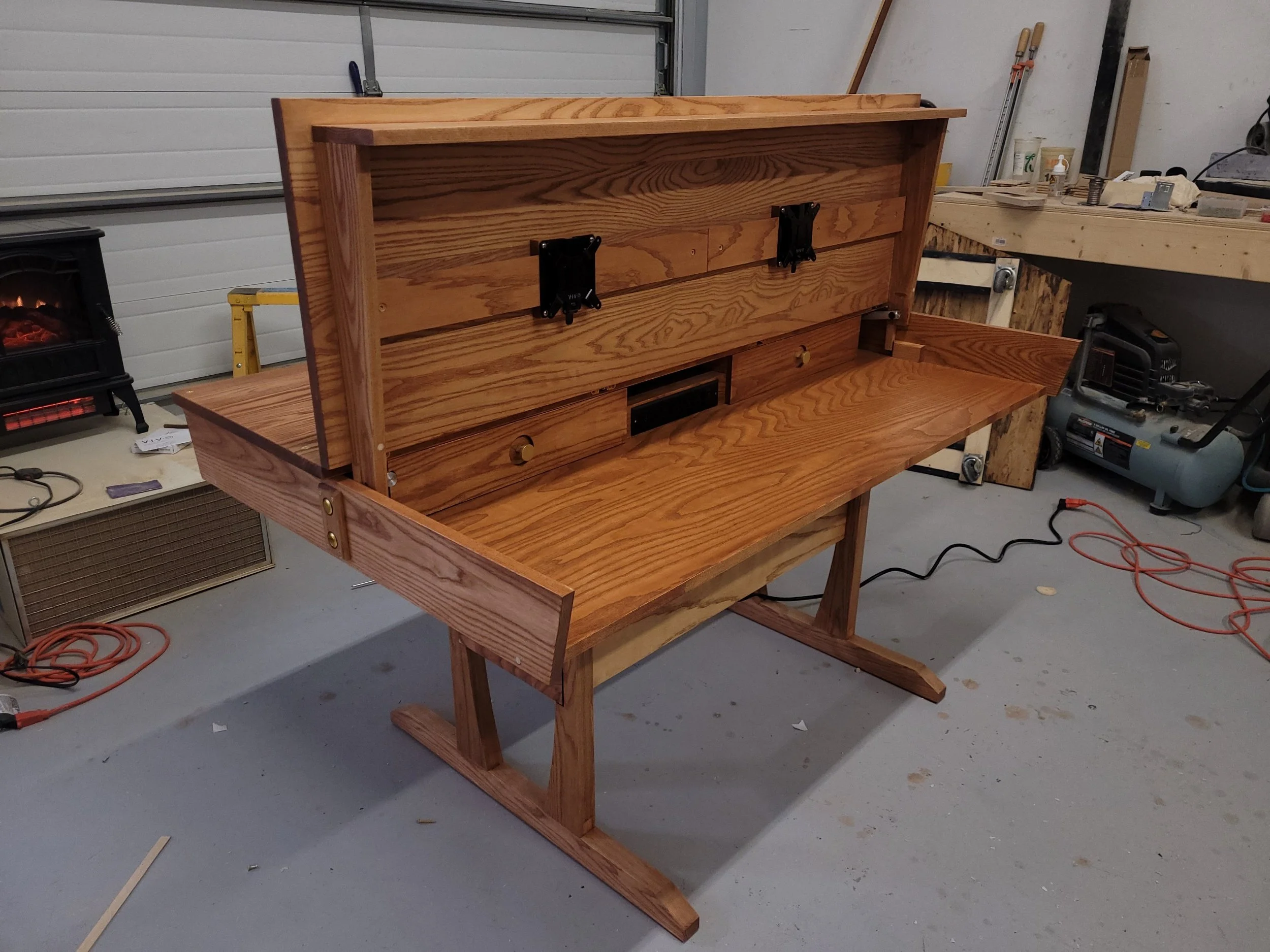 A partially assembled wooden desk with a raised back panel in a workshop, surrounded by tools and equipment.