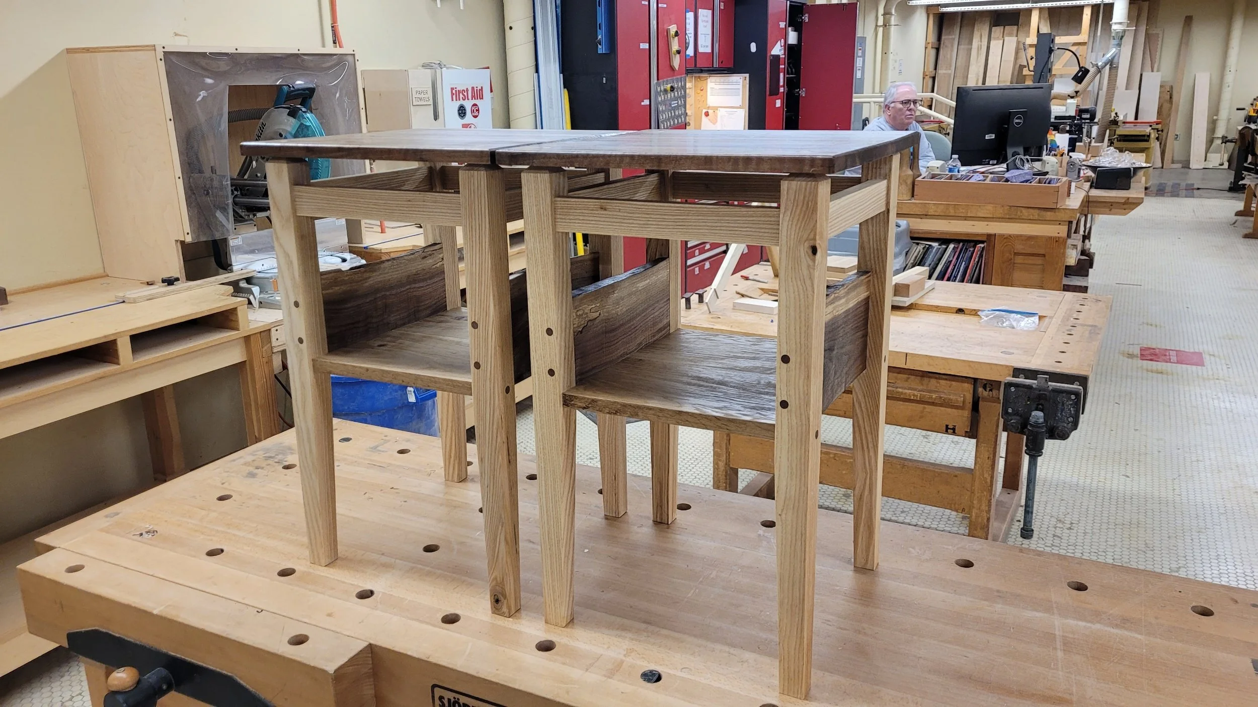 A work in progress wooden table on a workbench in a woodworking shop, with tools and equipment visible in the background.