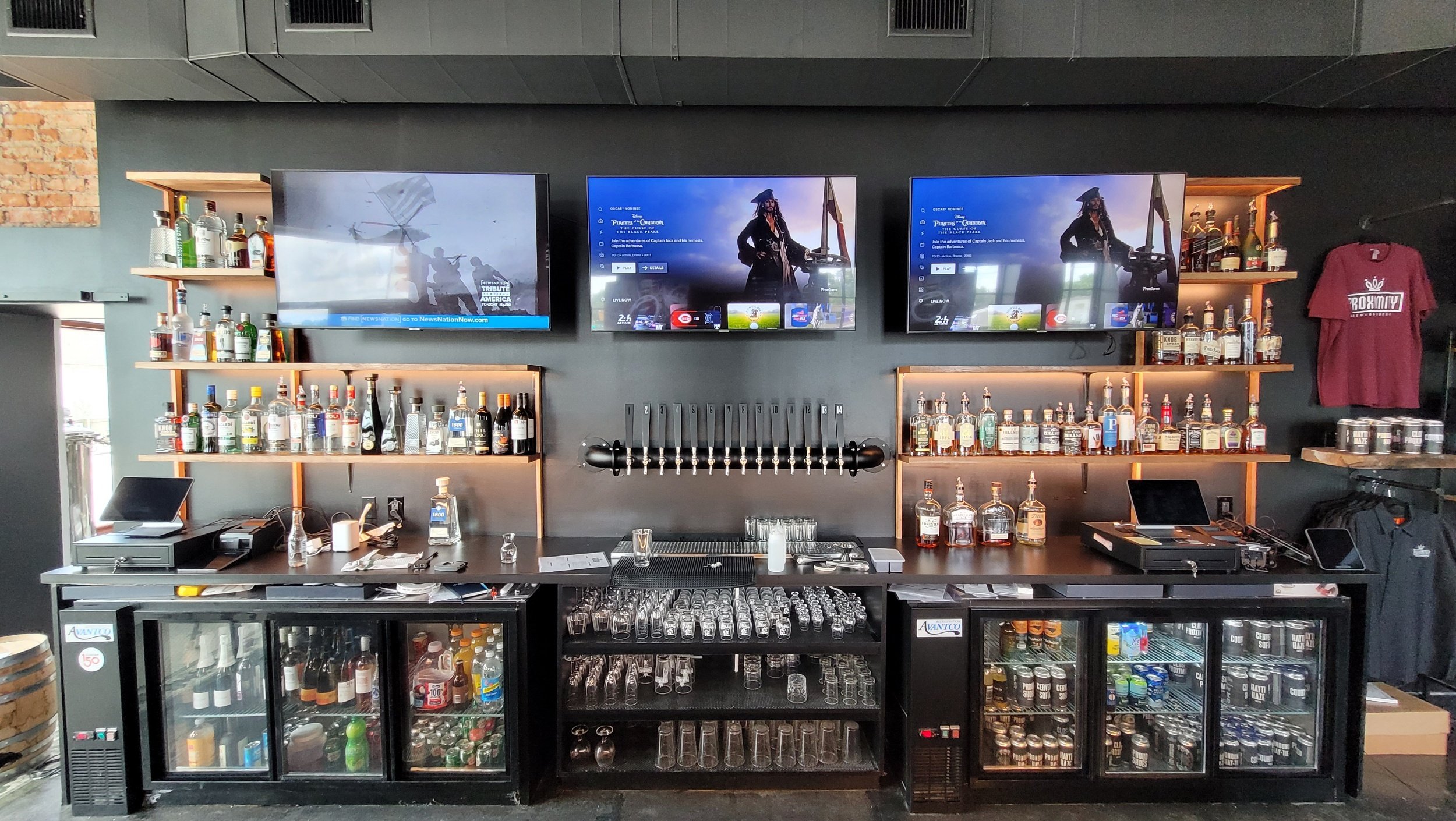 Bar area with shelves of liquor bottles, two beer taps, and a refrigerated display case containing various beverages, with three mounted TVs showing a man in a pirate costume and a helicopter scene.