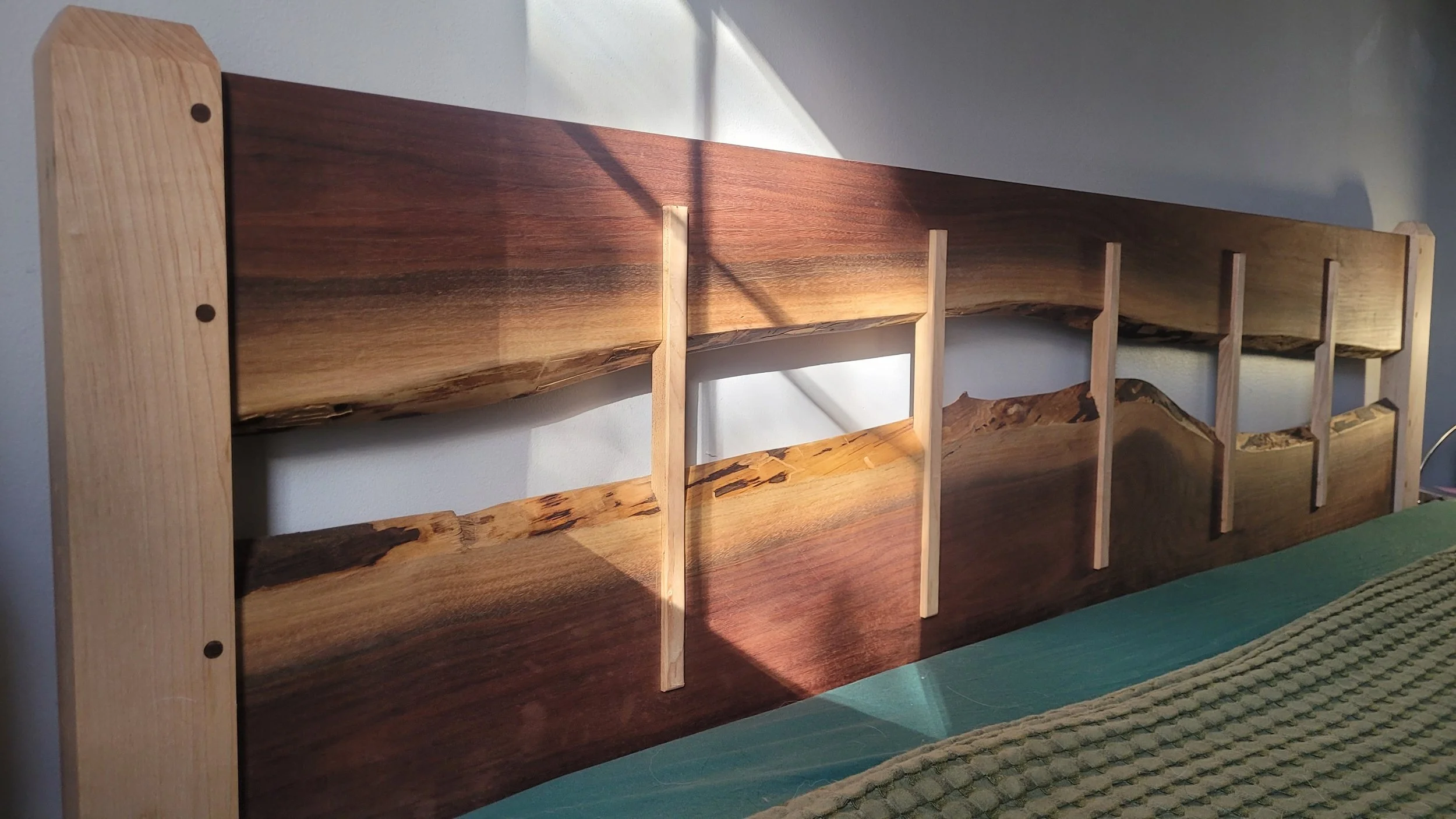 Close-up of a wooden headboard with a natural, live edge design, featuring horizontal and vertical wooden slats, mounted on a wall with sunlight and shadow.