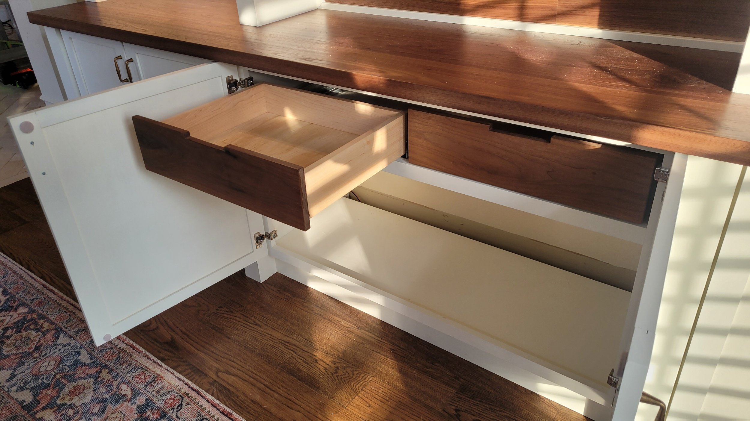 Open white cabinet with three drawers, one of which is partially pulled out, with a wood countertop and hardwood flooring underneath.