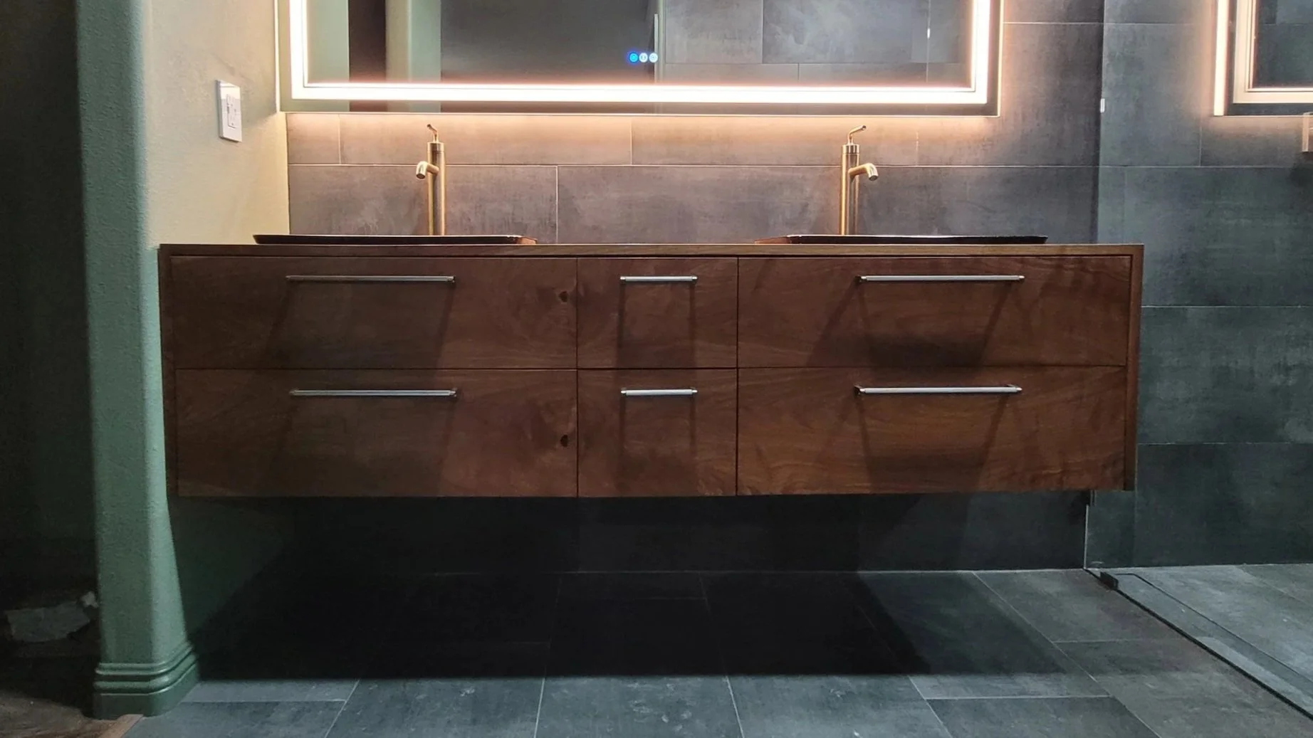 Modern bathroom vanity with a wooden cabinet, two sinks, and gold faucets, illuminated by backlit mirrors, against a gray tiled wall.