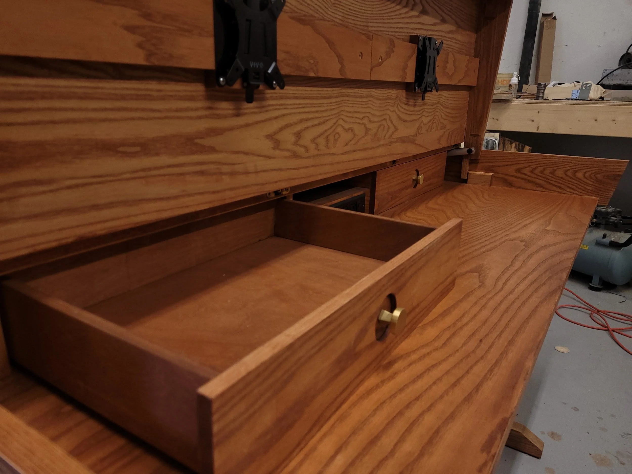 Unfinished wooden desk with open drawer in a workshop, with tools and equipment in the background.
