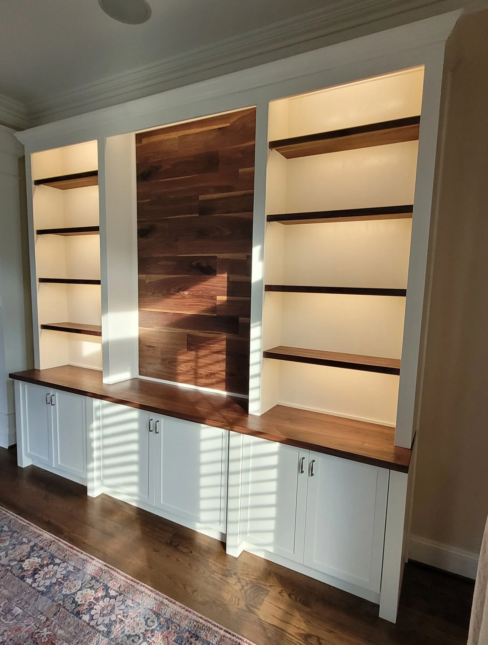 Built-in white bookshelf with dark wood shelves and accents, illuminated from above, with white cabinets at the bottom, a wood floor, and a patterned rug nearby.