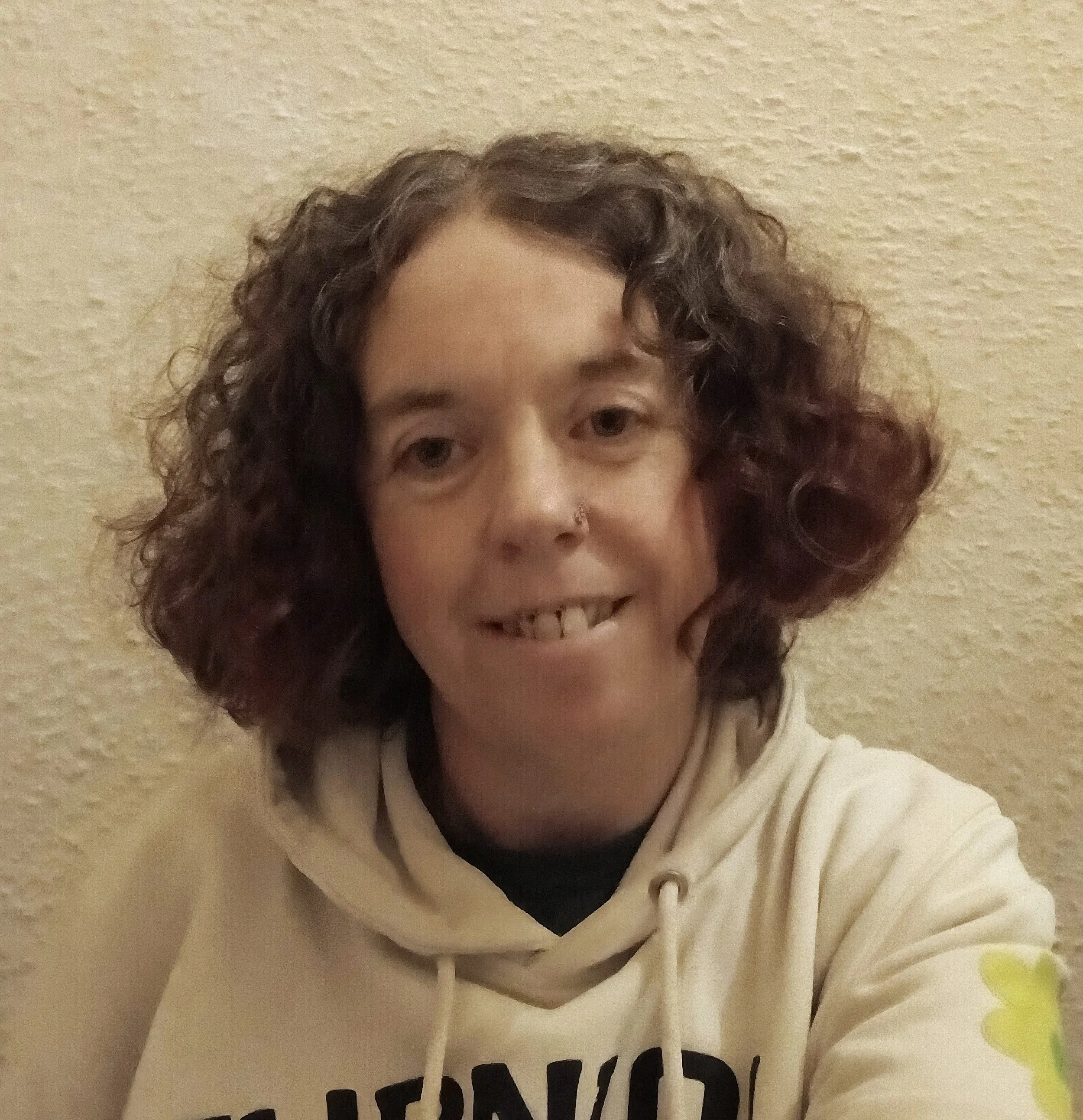 A person with curly brown hair taking a selfie against a beige textured wall, wearing a cream hoodie with black text.