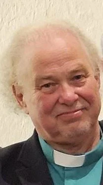 Photo of an elderly man with white hair, wearing a clerical collar and dark jacket, smiling slightly.