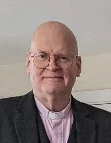 A man with glasses and a bald head wearing a clerical collar and dark blazer, smiling.
