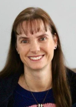 A woman with straight brown hair and bangs, smiling, wearing a navy blue top and small earrings.