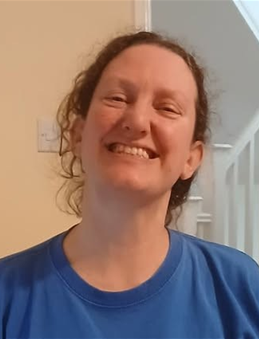 A woman with curly hair smiling in a blue shirt, standing indoors near a staircase.
