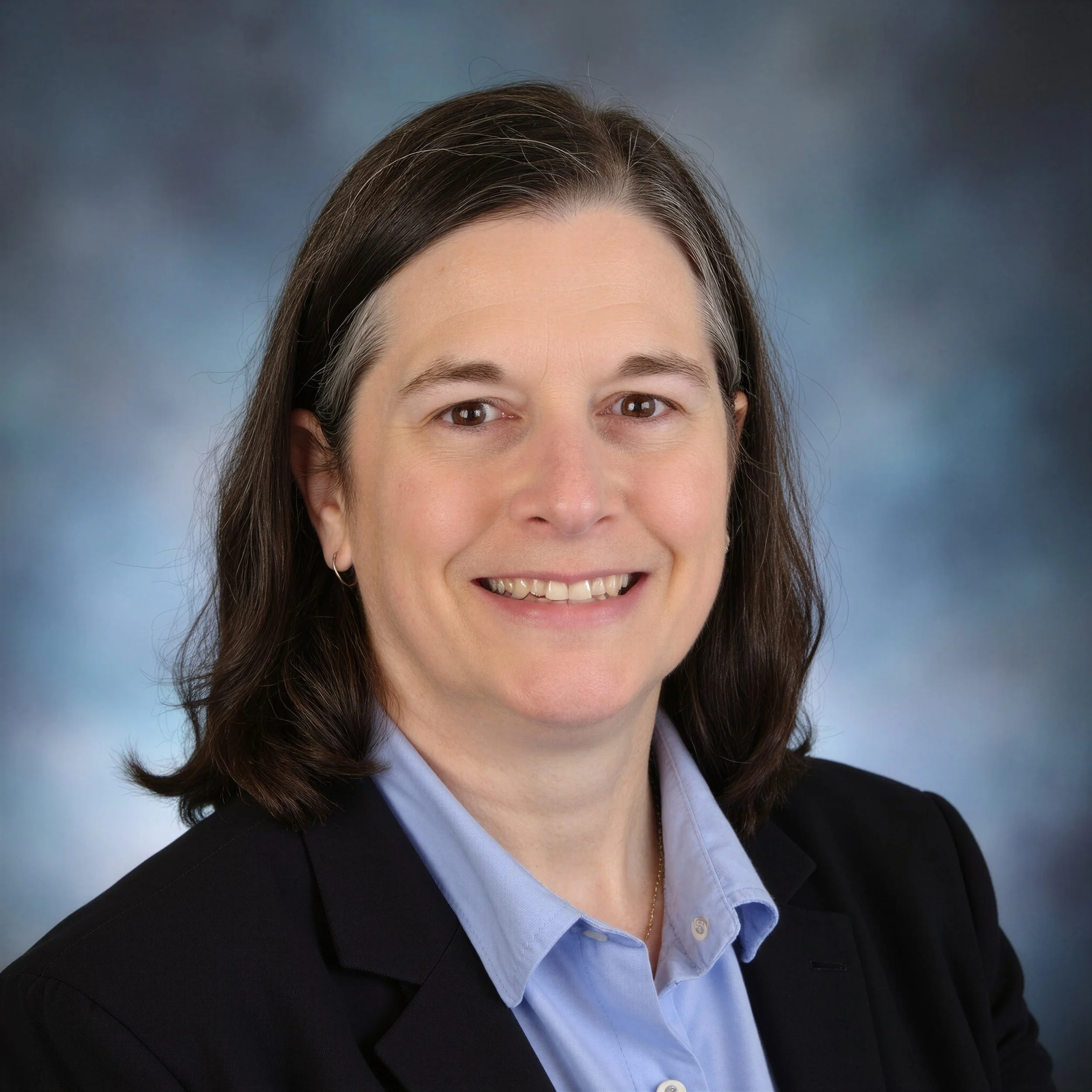 Close-up of a woman with brown hair, smiling, wearing a dark blazer over a light blue collared shirt, against a blurred blue-gray background.