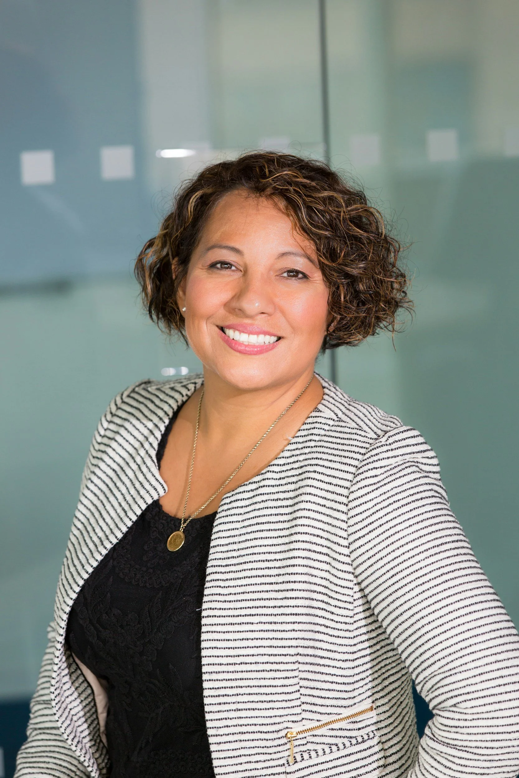Portrait of a woman with curly brown hair smiling, wearing a black lace top, striped blazer, and gold jewelry, standing in front of a glass wall.