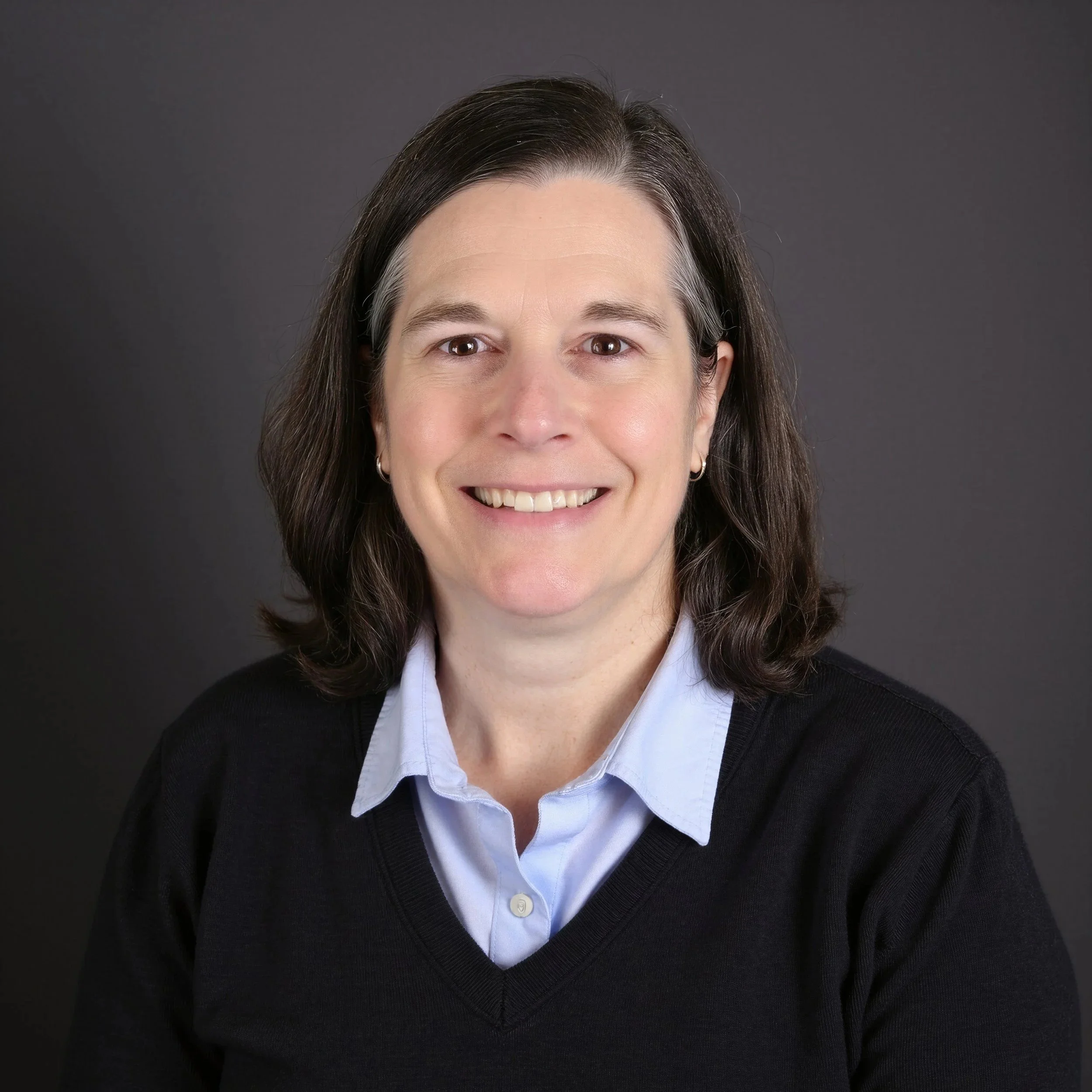 Close-up of a woman with dark brown hair, smiling, wearing a white collared shirt and a black cardigan against a dark gray background.