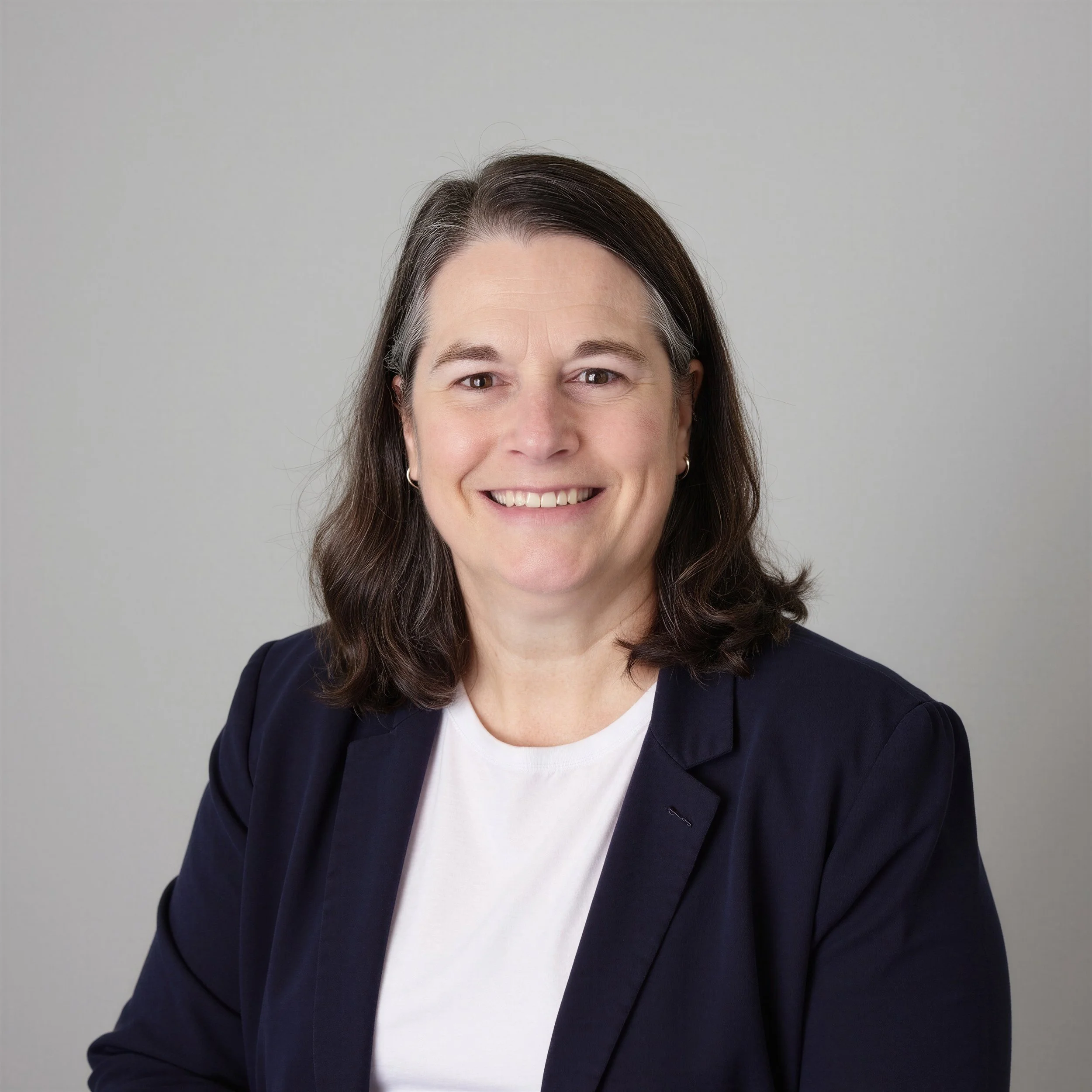 Close-up of a woman with shoulder-length brown hair, smiling, wearing a navy blazer over a white shirt, against a plain gray background.