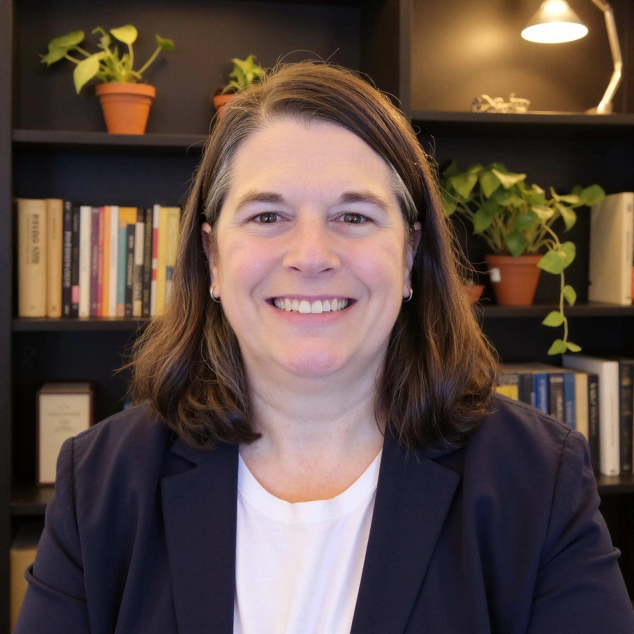 Close-up of a smiling woman with dark brown hair and earrings, wearing a black blazer and white shirt, in front of a dark bookshelf with books and potted plants in a well-lit room.