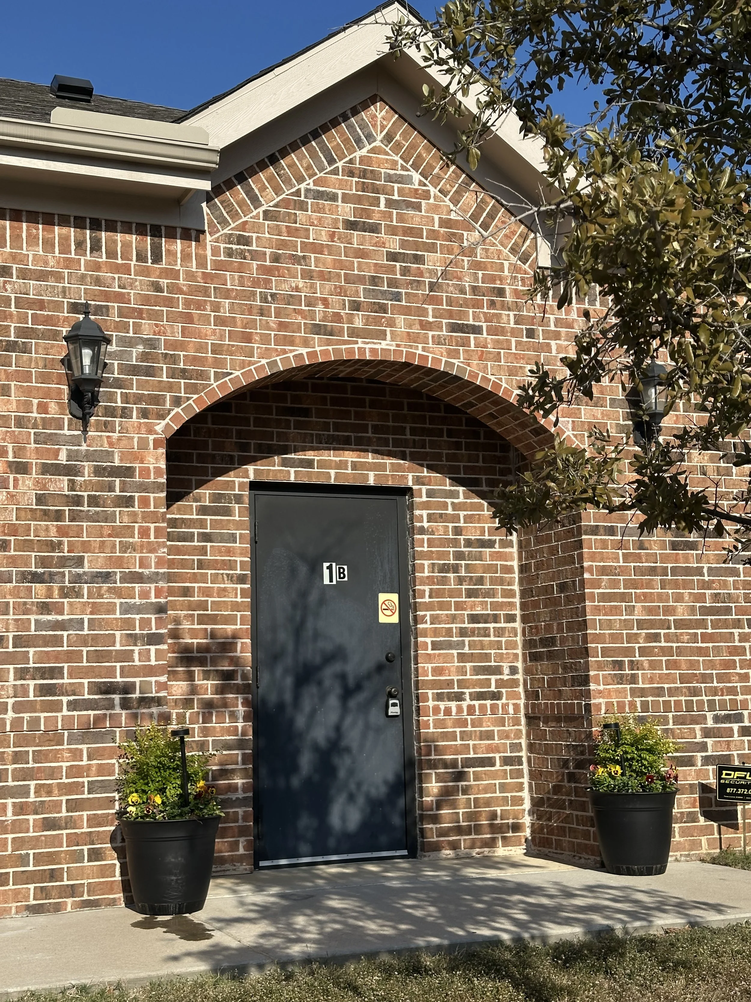 Brick building entrance with a dark blue door labeled '1B', flanked by lantern-style wall lights and two potted plants. A small sign is visible in the grass.
