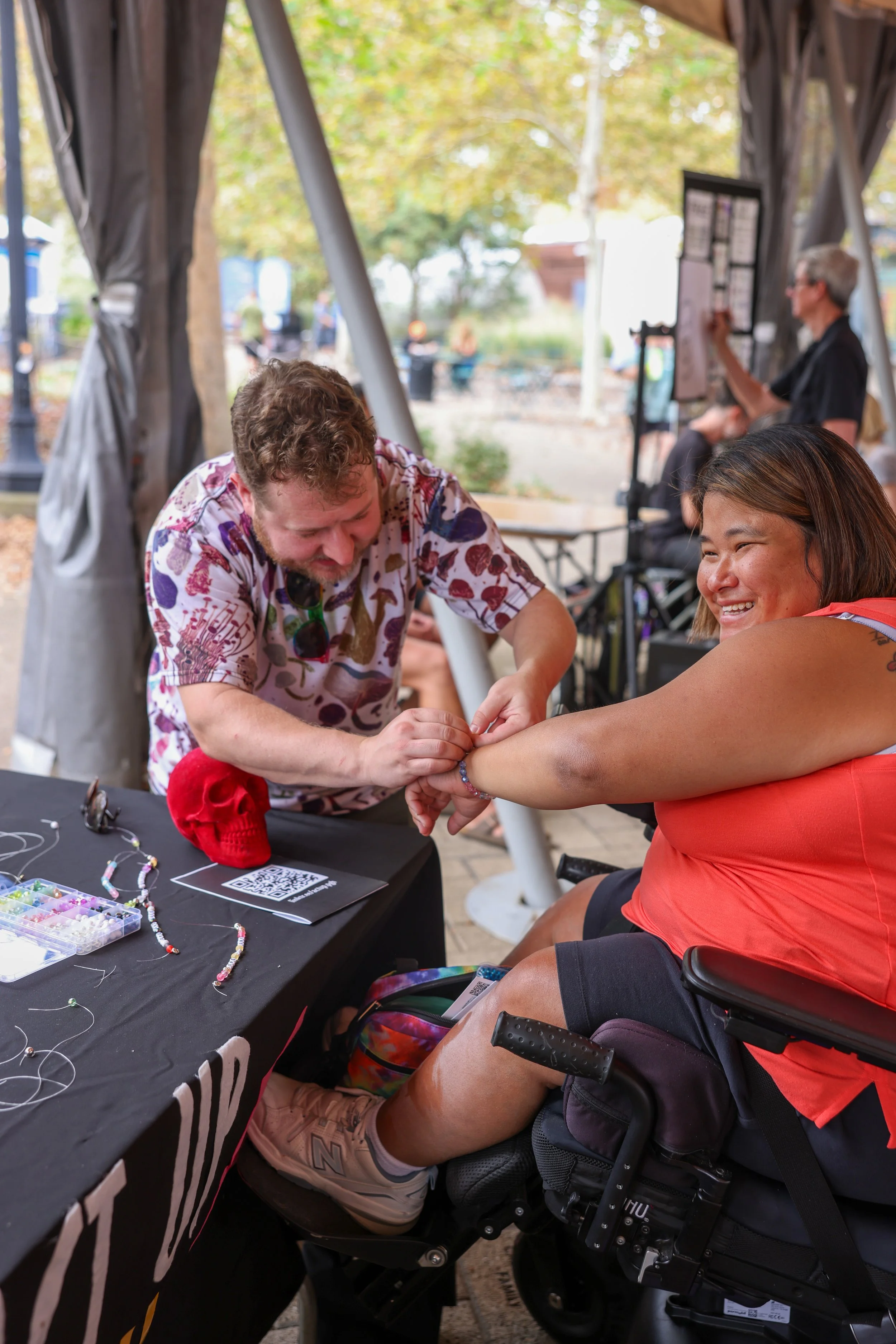 Asian woman in coral top receives friendship bracelet from white man in patterned shirt