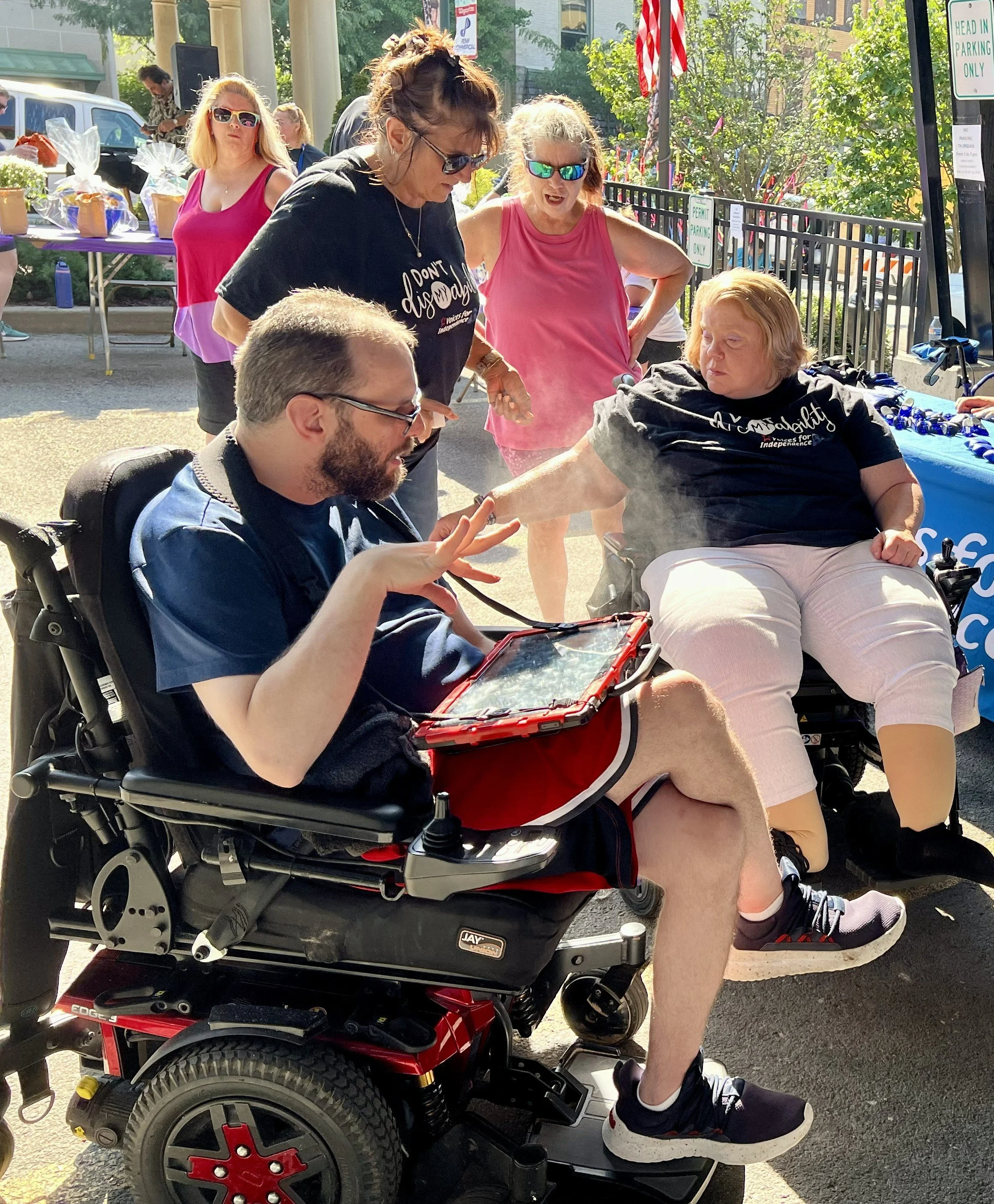 A man with glasses using a power chair and communication board chats with a woman with red hair who is also a power chair user.