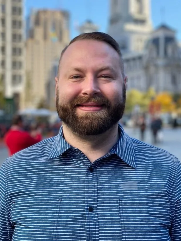 Smiling white man with a mustache and beard wearing a blue shirt with a city in the background