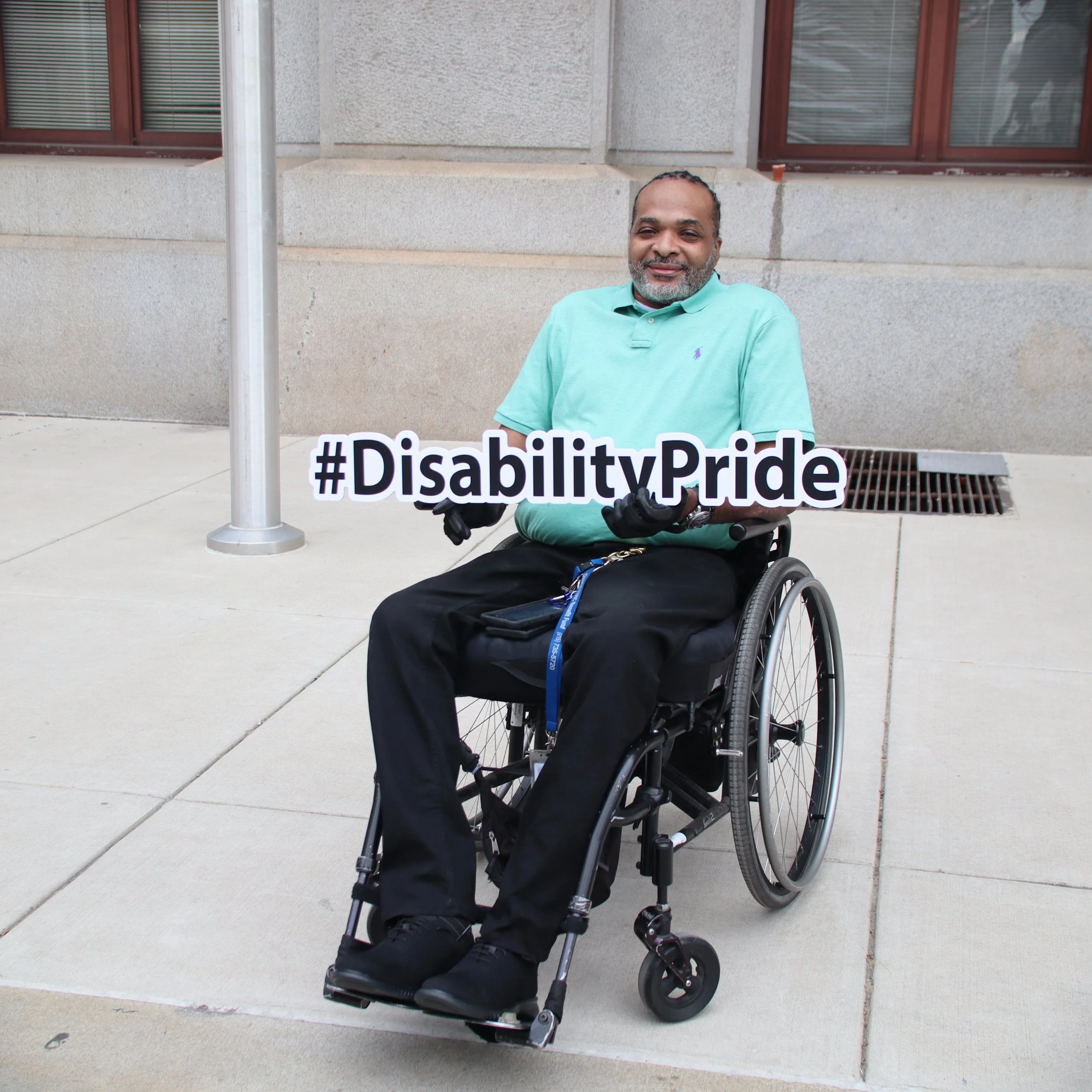 Black wheelchair user holding up white cutout with #DisabilityPride