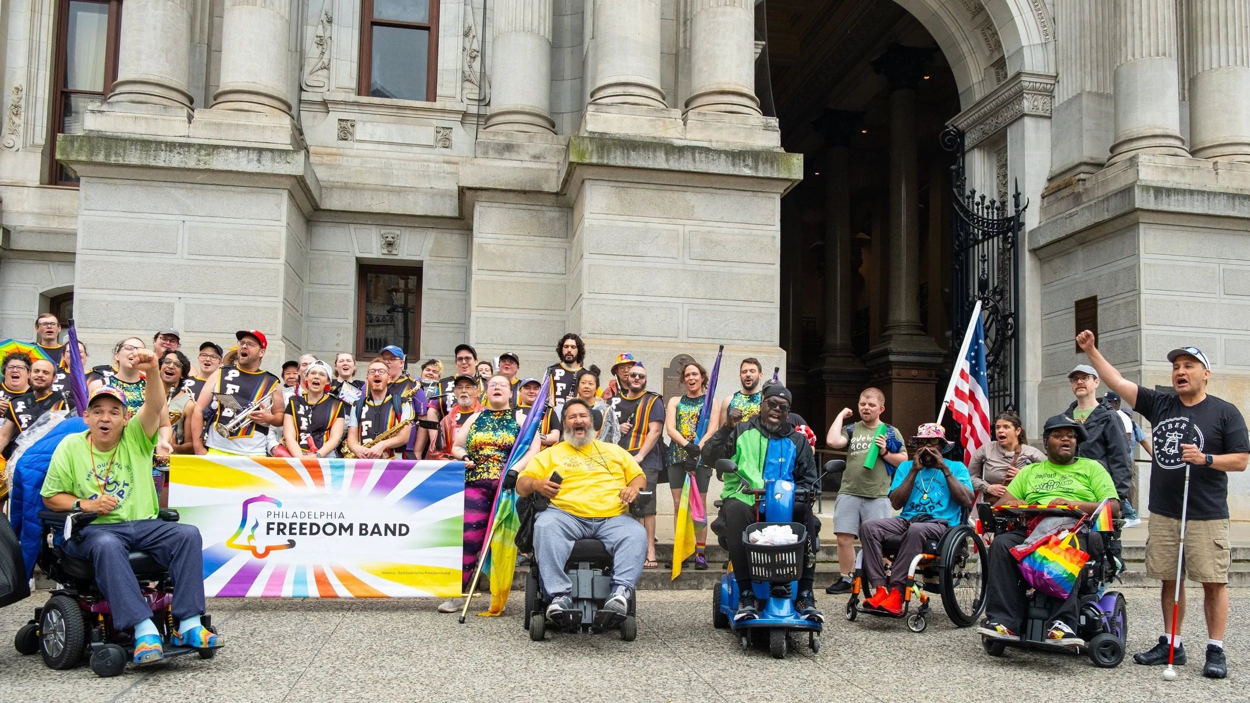 A diverse group of disabled and nondisabled people gather at Disability Pride Philly. Several people use power wheelchairs and mobility devices. They hold rainbow flags, an American flag, and a colorful banner reading "Philadelphia Freedom Band."