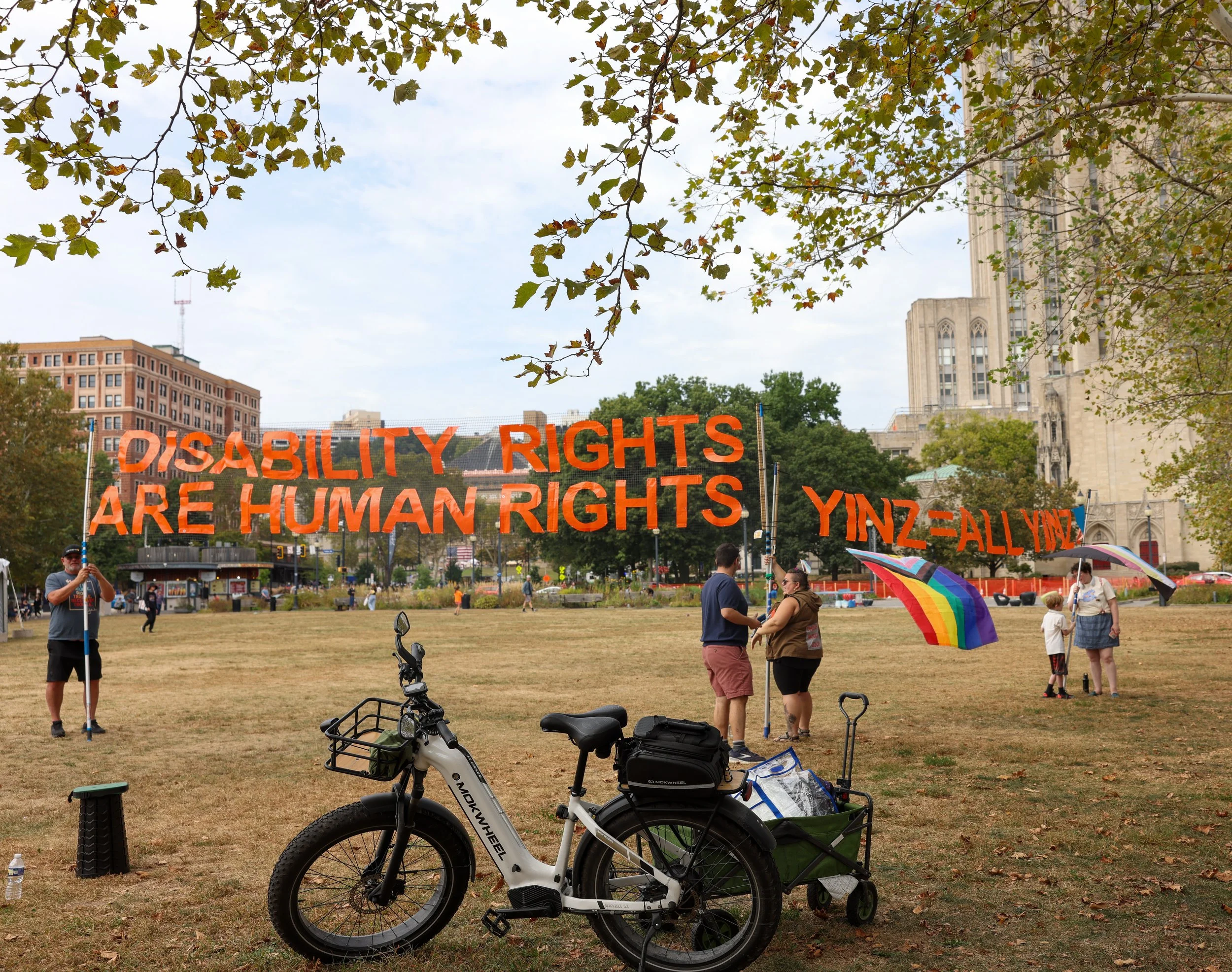 Behind a white e-bike are people setting up a banner saying, "Disability Rights Are Human Rights." To the right, someone holds a pride flag and another banner says "Yinz = All Yinz.