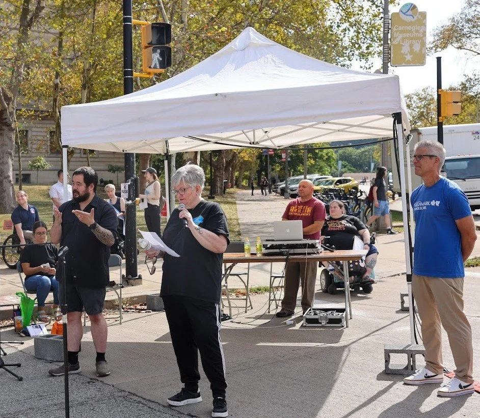 Vicki Landers, wearing a black shirt and glasses, speaks into a microphone at an outdoor event under a white tent. Community members and audio equipment are visible in the background.