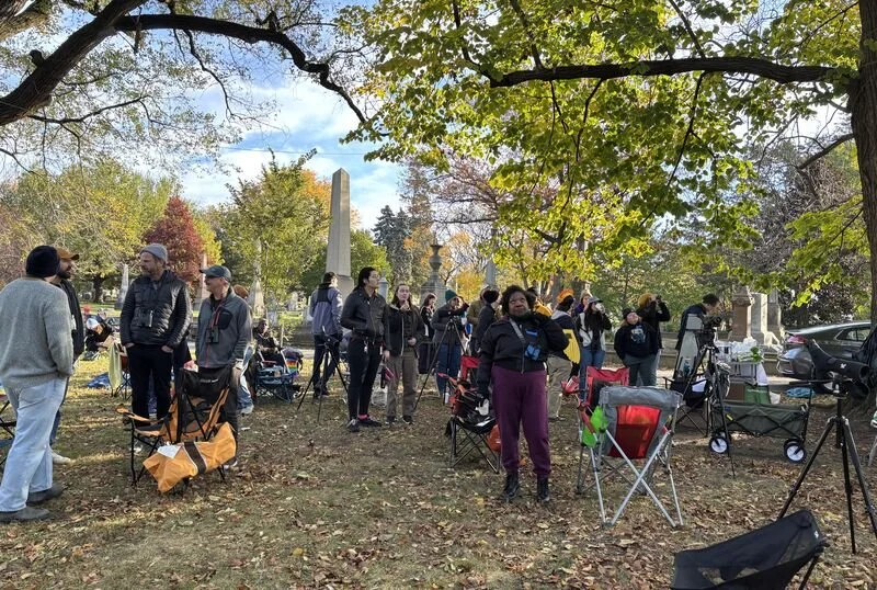 Group of people in a wooded cemetery area with chairs, wagons, and bird watching gear.
