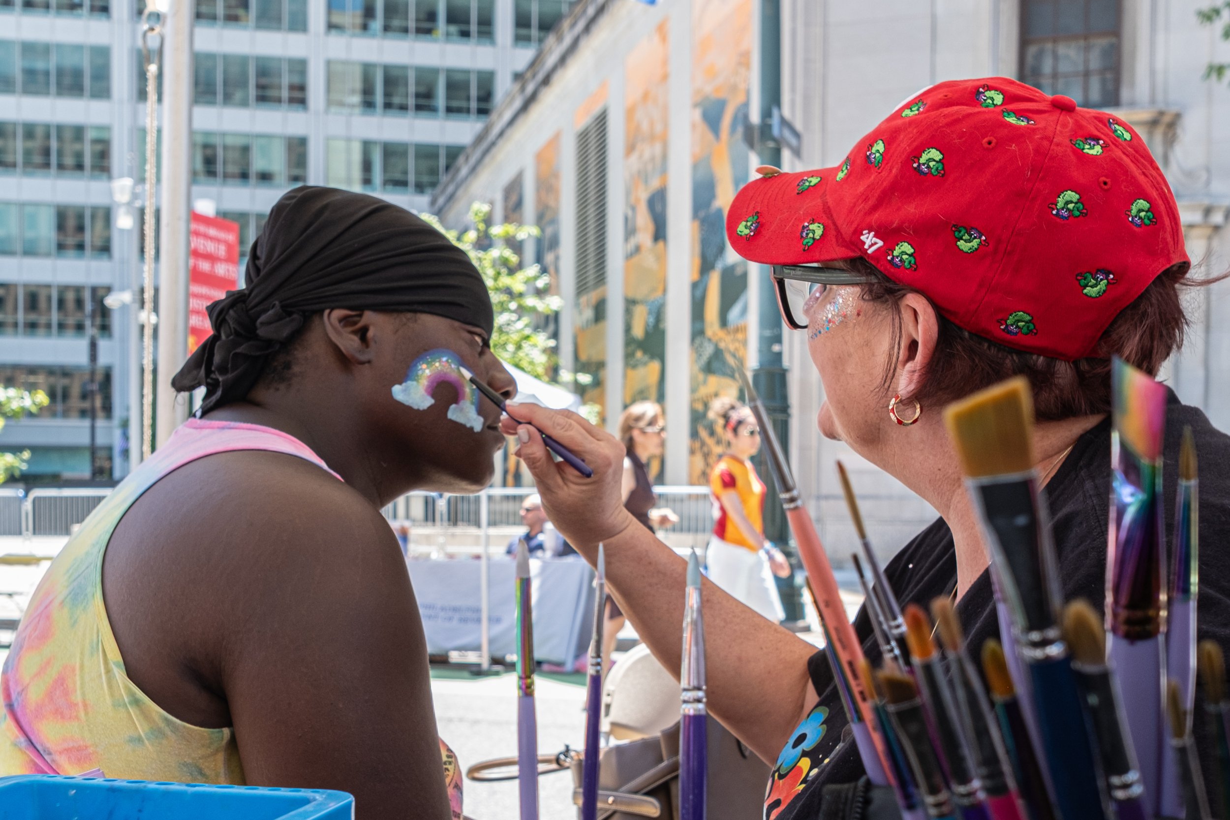 Face painter in a red hat is painting a rainbow on a man's face.