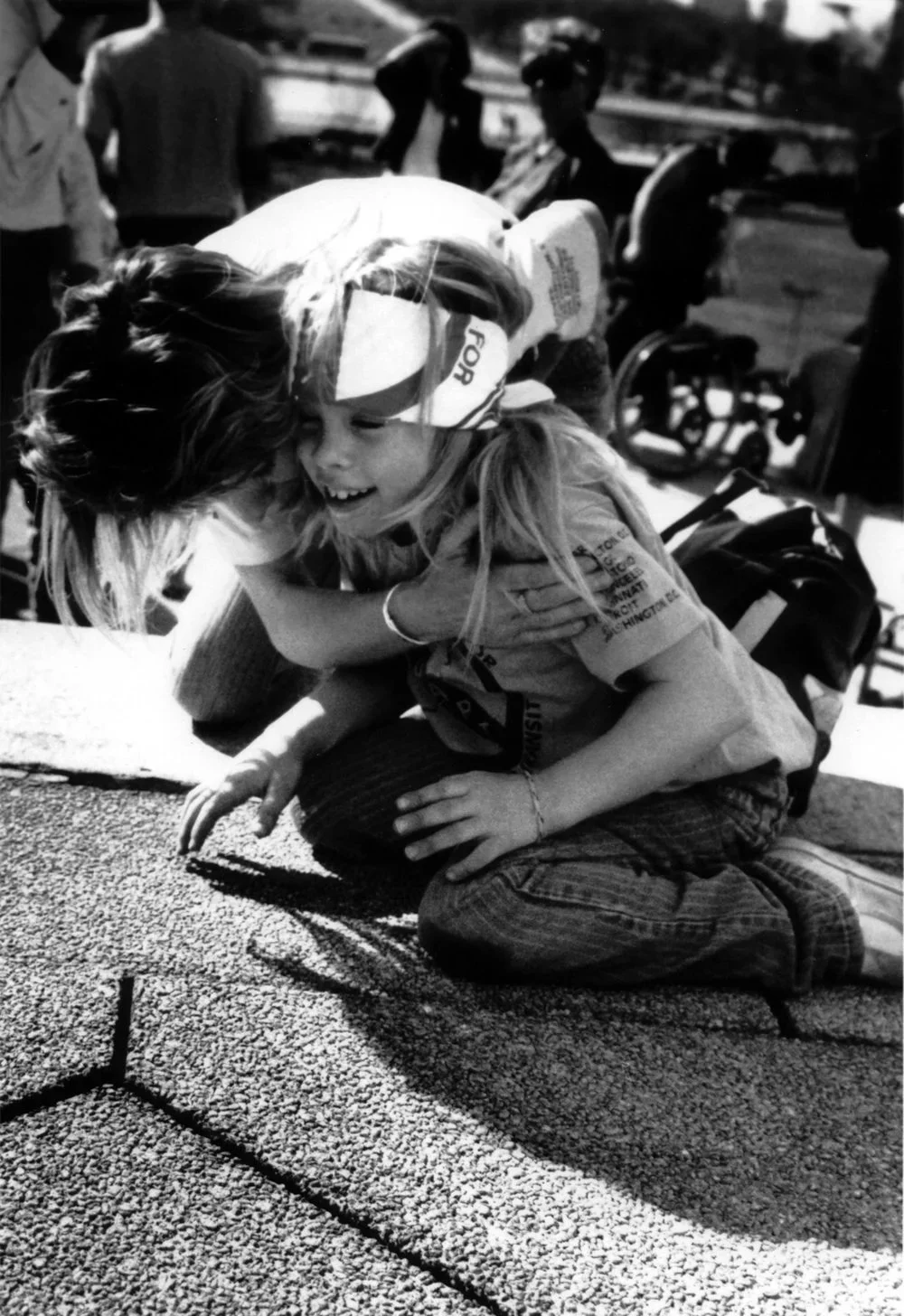 A small blond child kneels at the top of a granite stair case and is hugged by an adult. She is smiling and wearing an ADAPT bandana.