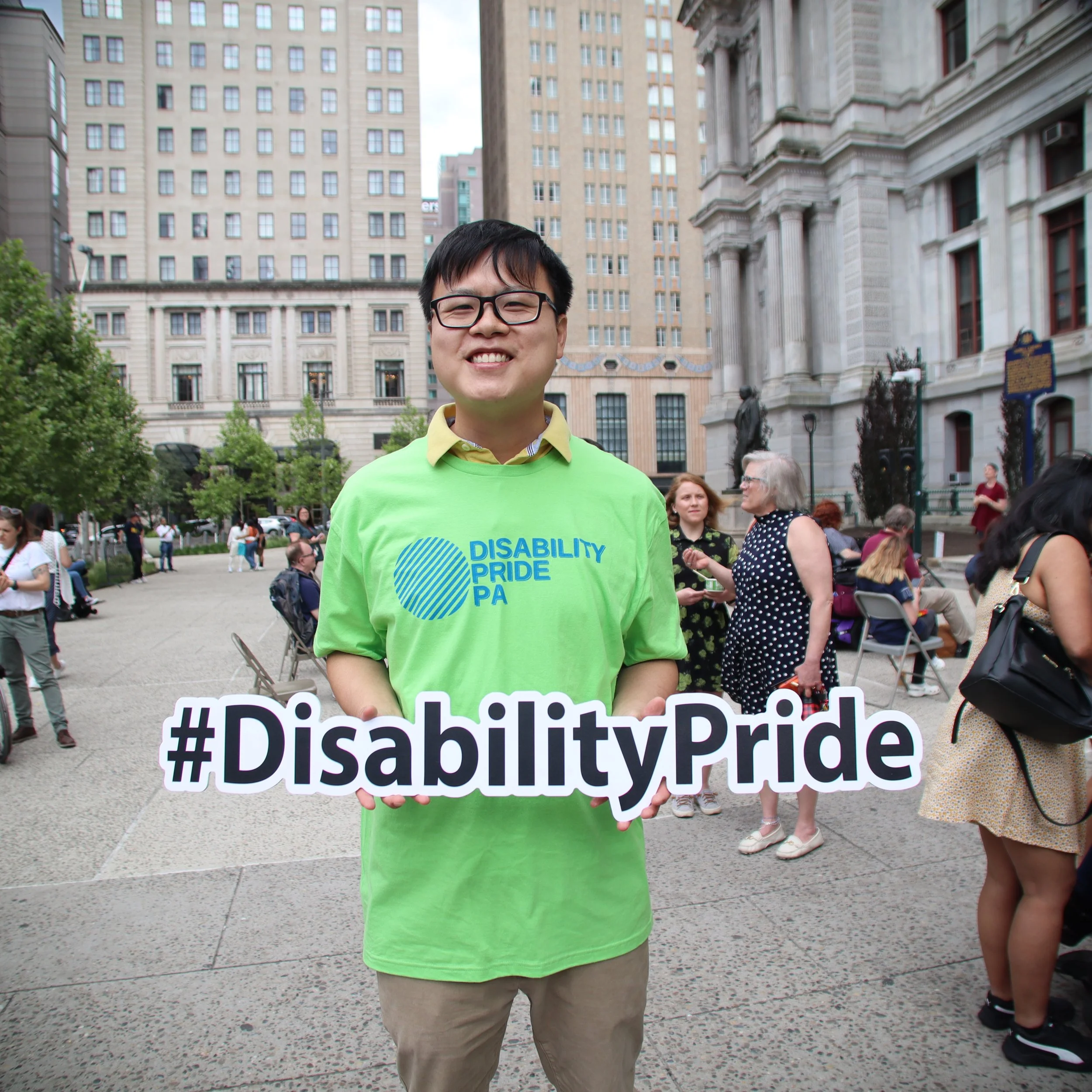 Asian American man with glasses wearing a bright green Disability Pride PA shirt and holding #DisabilityPride sign