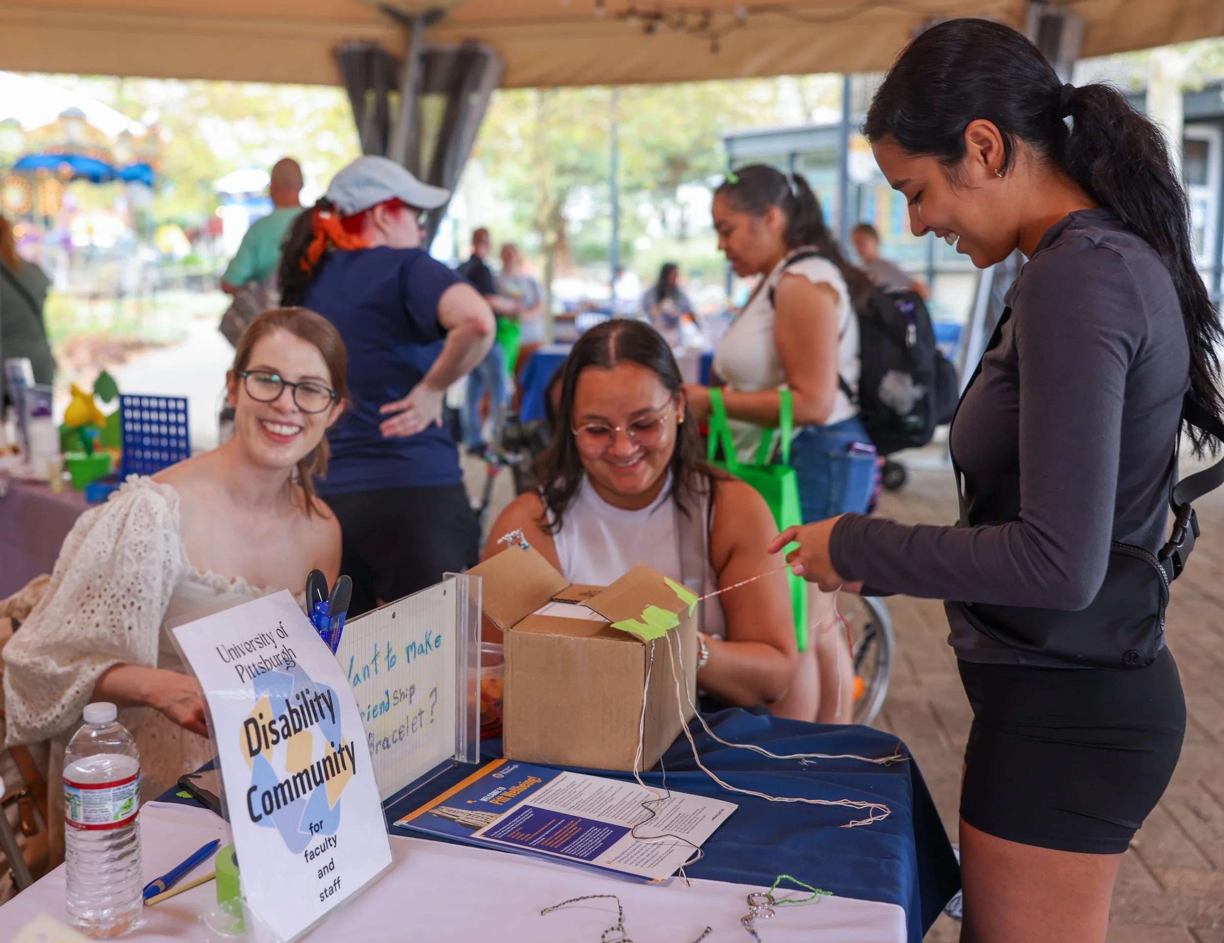 Three women at University of Pittsburgh Disability Community table, attendee making friendship bracelet
