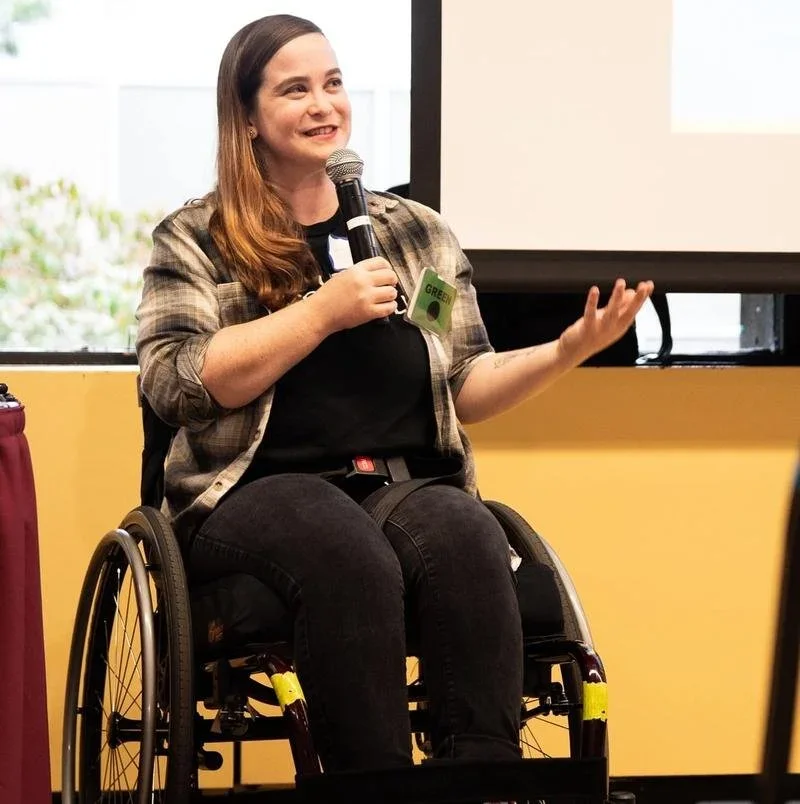 White wheelchair user with long hair speaking in a microphone.