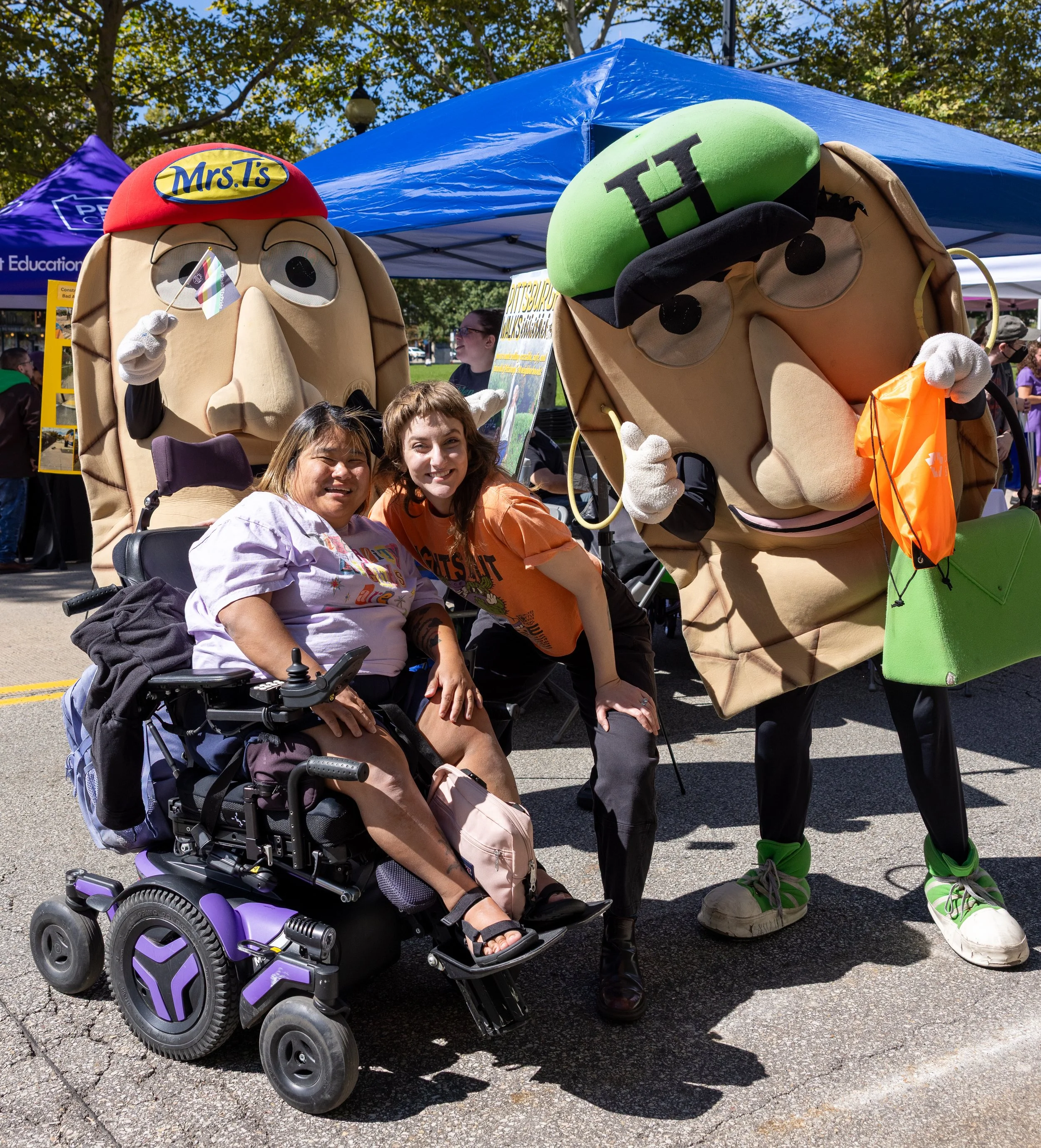Libby in her purple wheelchair smiles with Mallory leaned down near her and two Perogies mascots from the Pittsburgh Pirates!