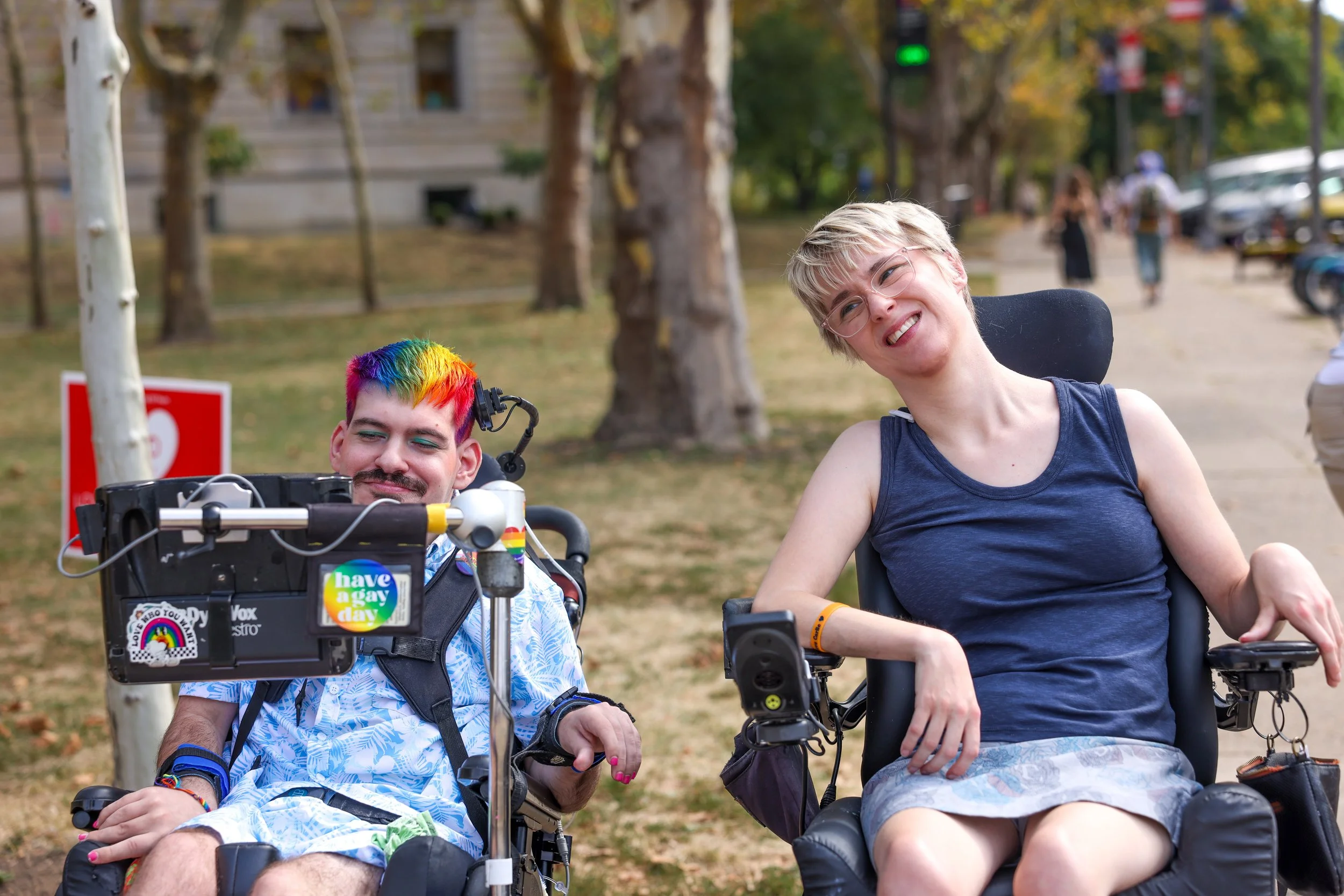 Two power chair users smiling, one with rainbow-dyed hair
