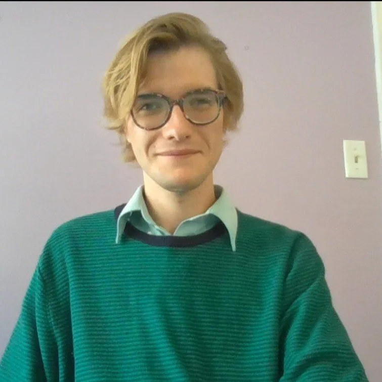Nico is a blonde'haired man with glasses. He is seated in front of a purple indoor wall smiling at the camera. He is wearing a blue collared shirt underneath a green sweater.