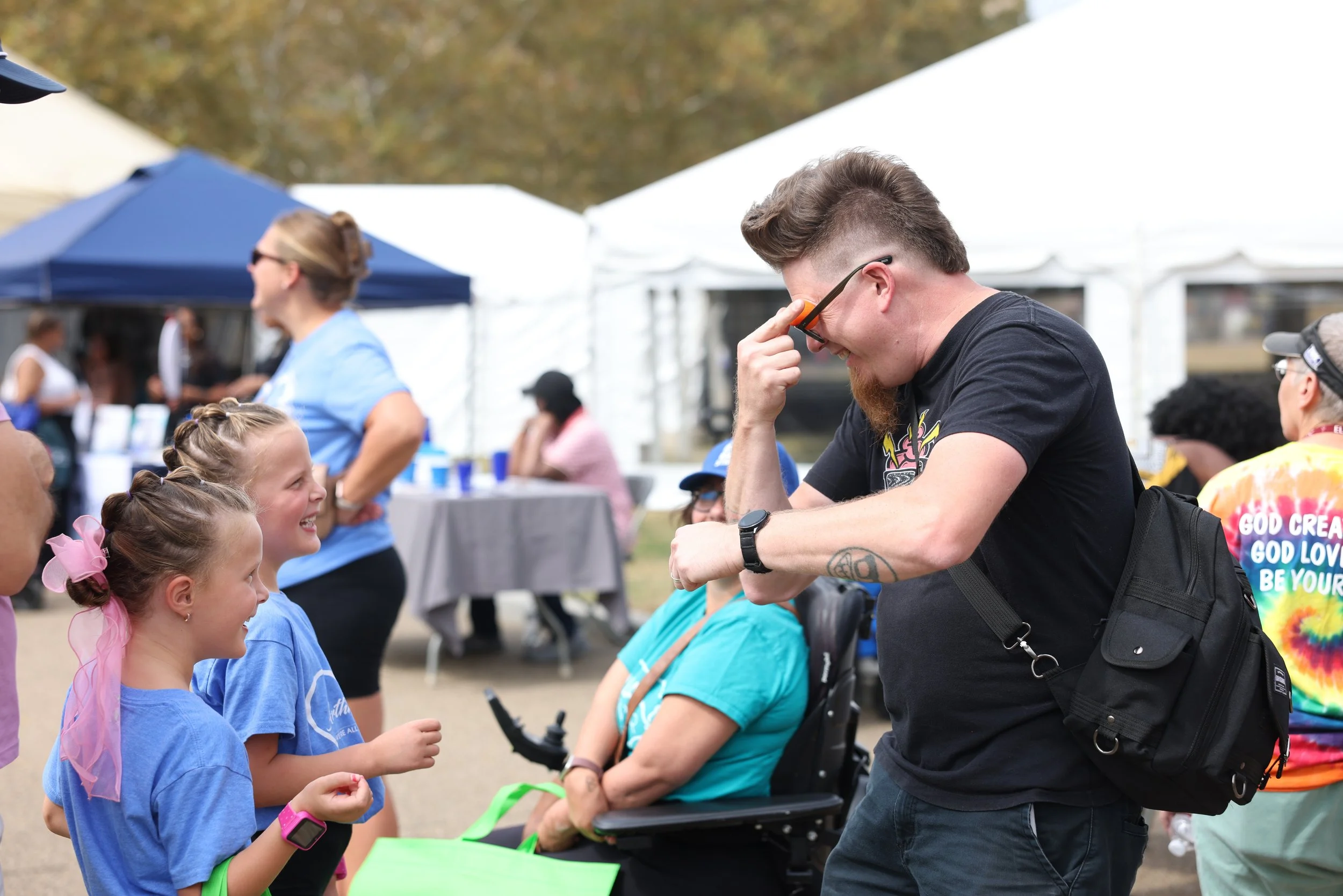 Two young white girls with braids watch a white man in black T-shirt goof off with sunglasses, all smiling