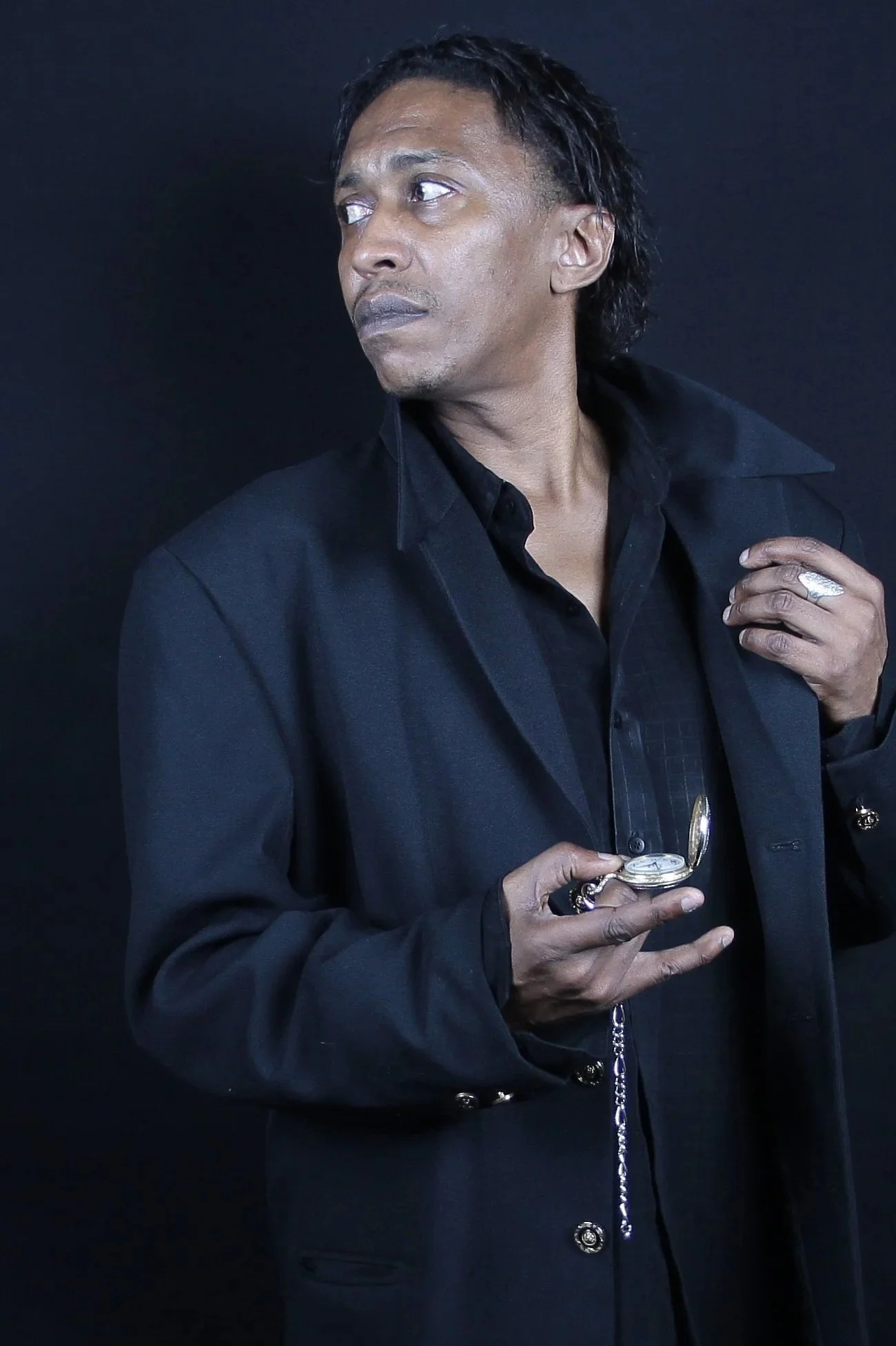 Black man dressed in black looking to the side and holding a time piece.
