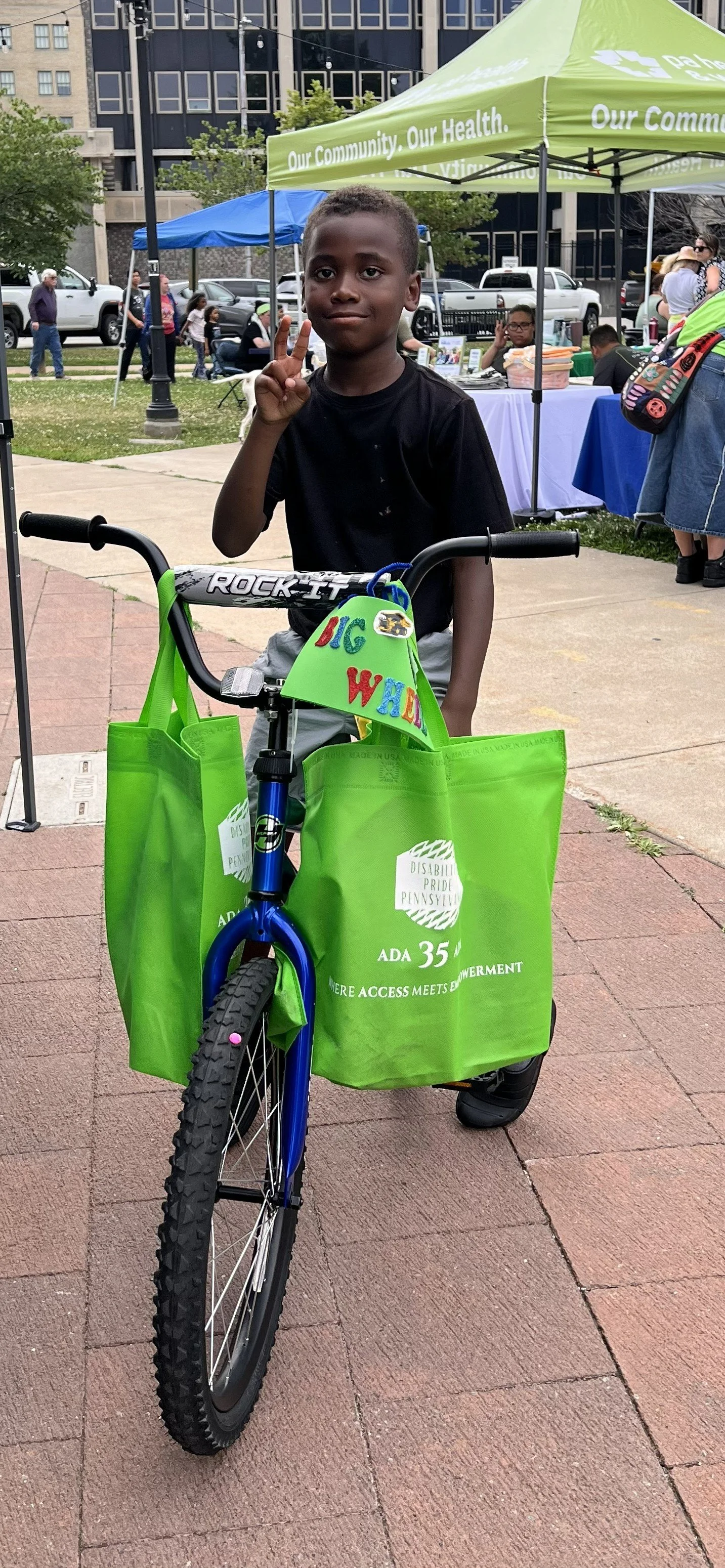 A Black boy gives a peace sign while sitting on his bike. Disability Pride bags hang from the handles and you can see vendor booths in the background.