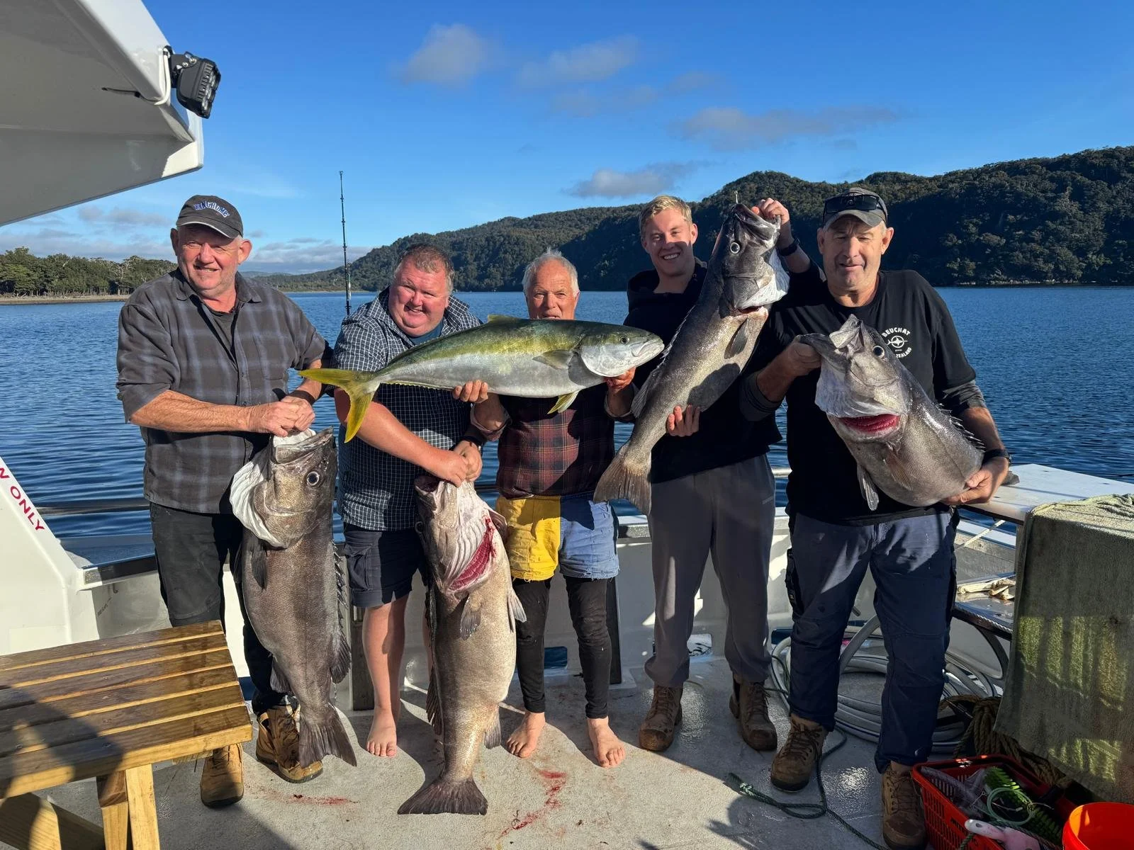 Five men on a boys trip aboard our boat holding large fish they caught, with a the beauty of Fiordland in the background.