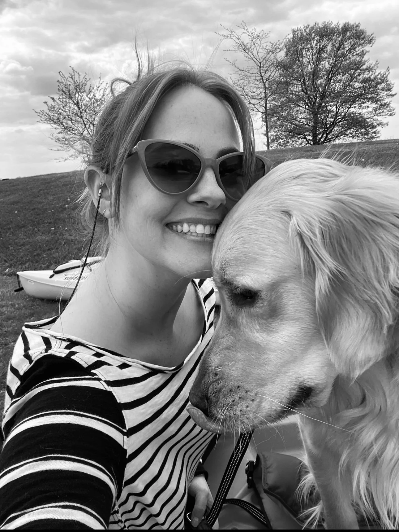 A woman smiling with sunglasses, taking a selfie with her golden retriever dog outdoors. There are trees and a boat in the background.