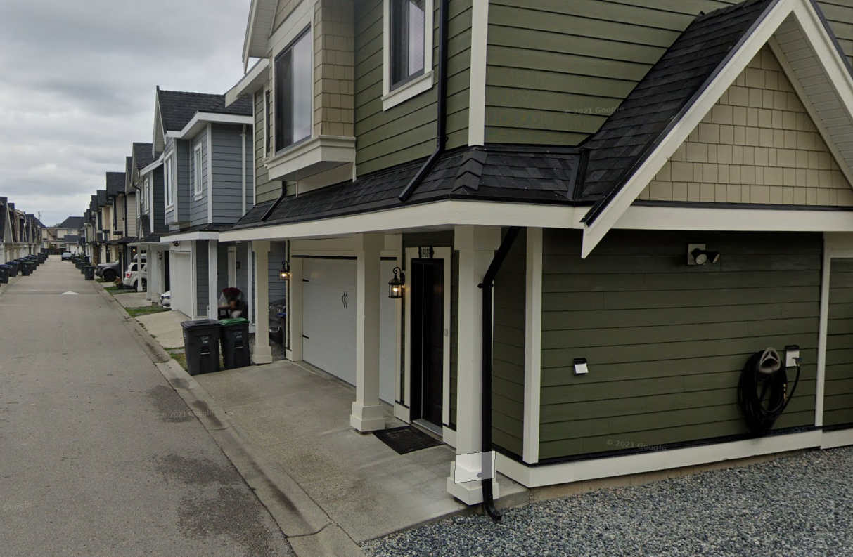 A row of modern townhouses with garages, a person working near a trash bin, and a gray street under cloudy skies.