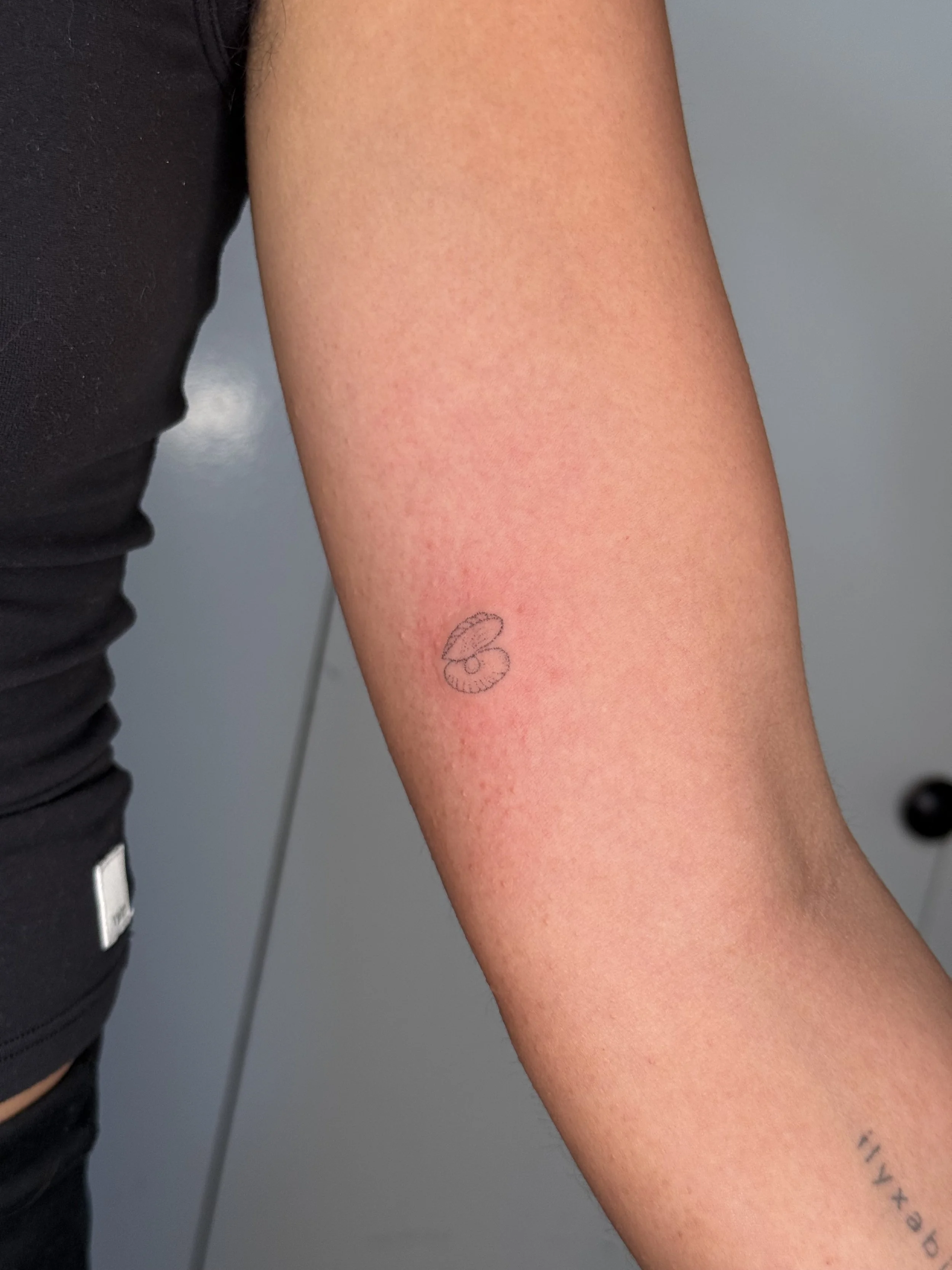 A close-up of a person's arm with light skin, showing a small tattoo near the wrist and a faint red mark on the skin. The background is a plain, gray surface.