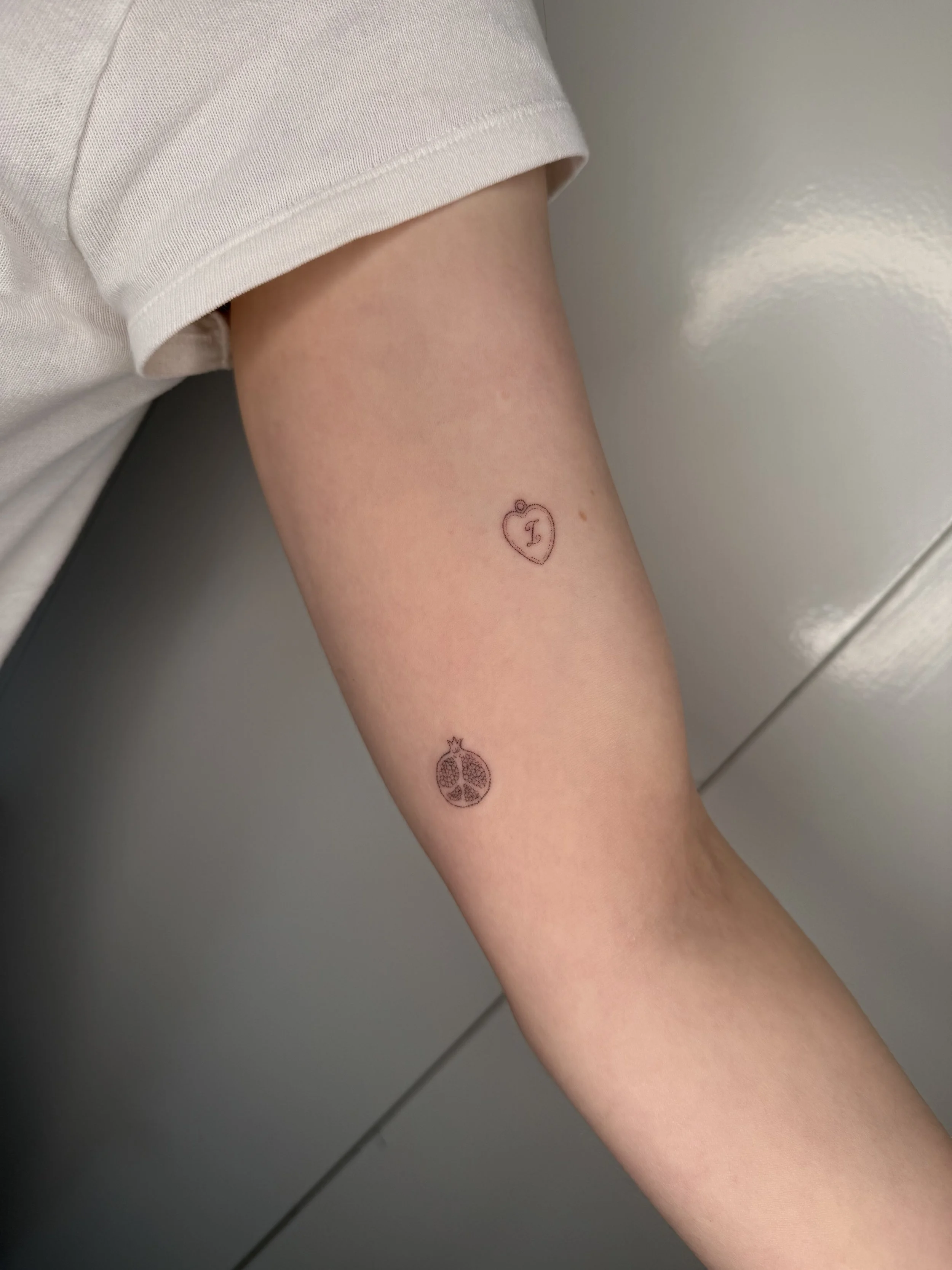 Close-up of a person's upper arm with three small tattoos: a heart with the letter 'L' inside, a pomegranate, and a pineapple.