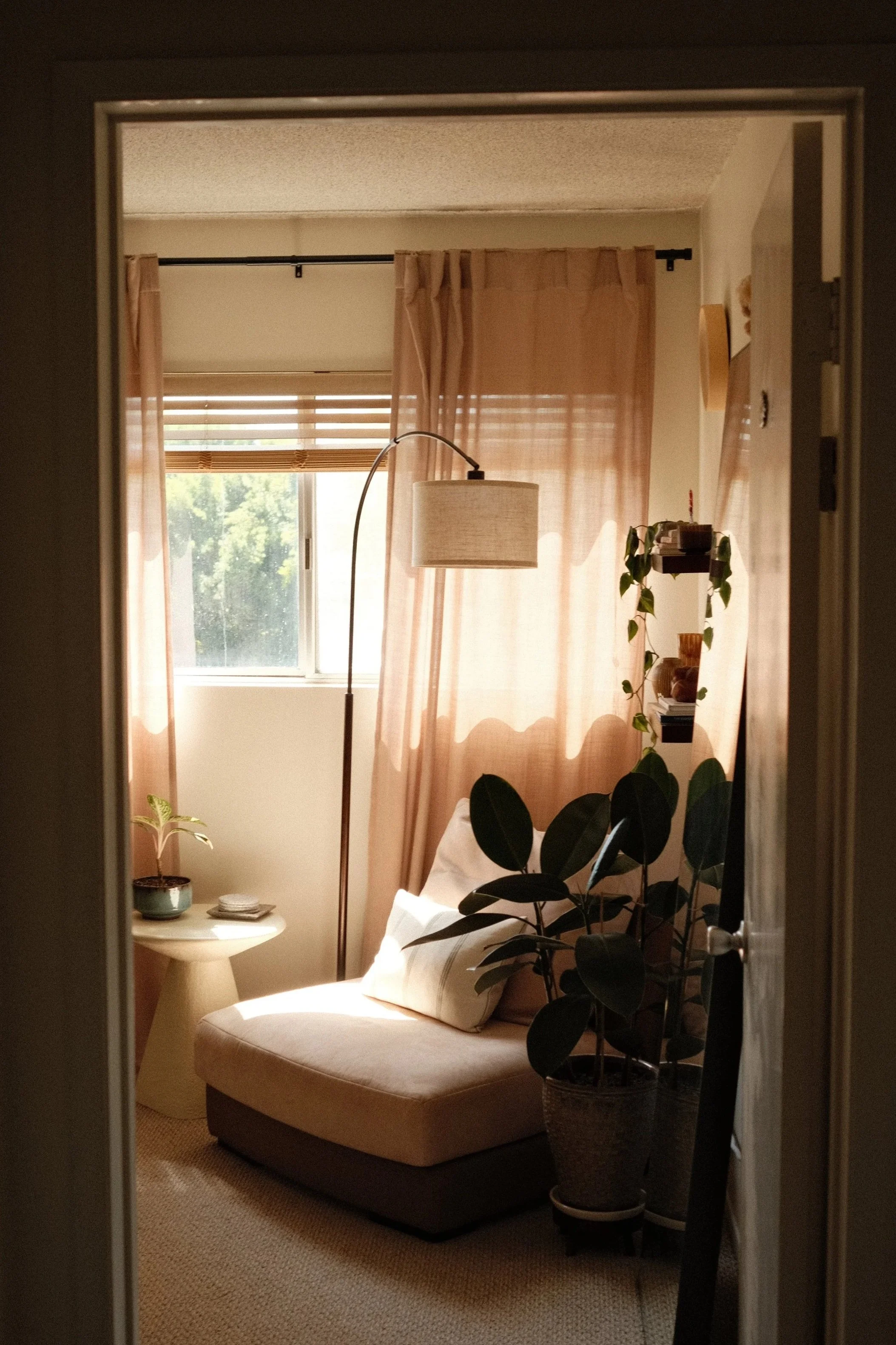 A cozy corner of a room with a beige chaise lounge, a white pillow, and a small side table with a potted plant and some items on it. There's a window with blinds and sheer curtains, and a tall floor lamp with a curved arm and a lampshade. A small shelf with a plant and books is also visible.