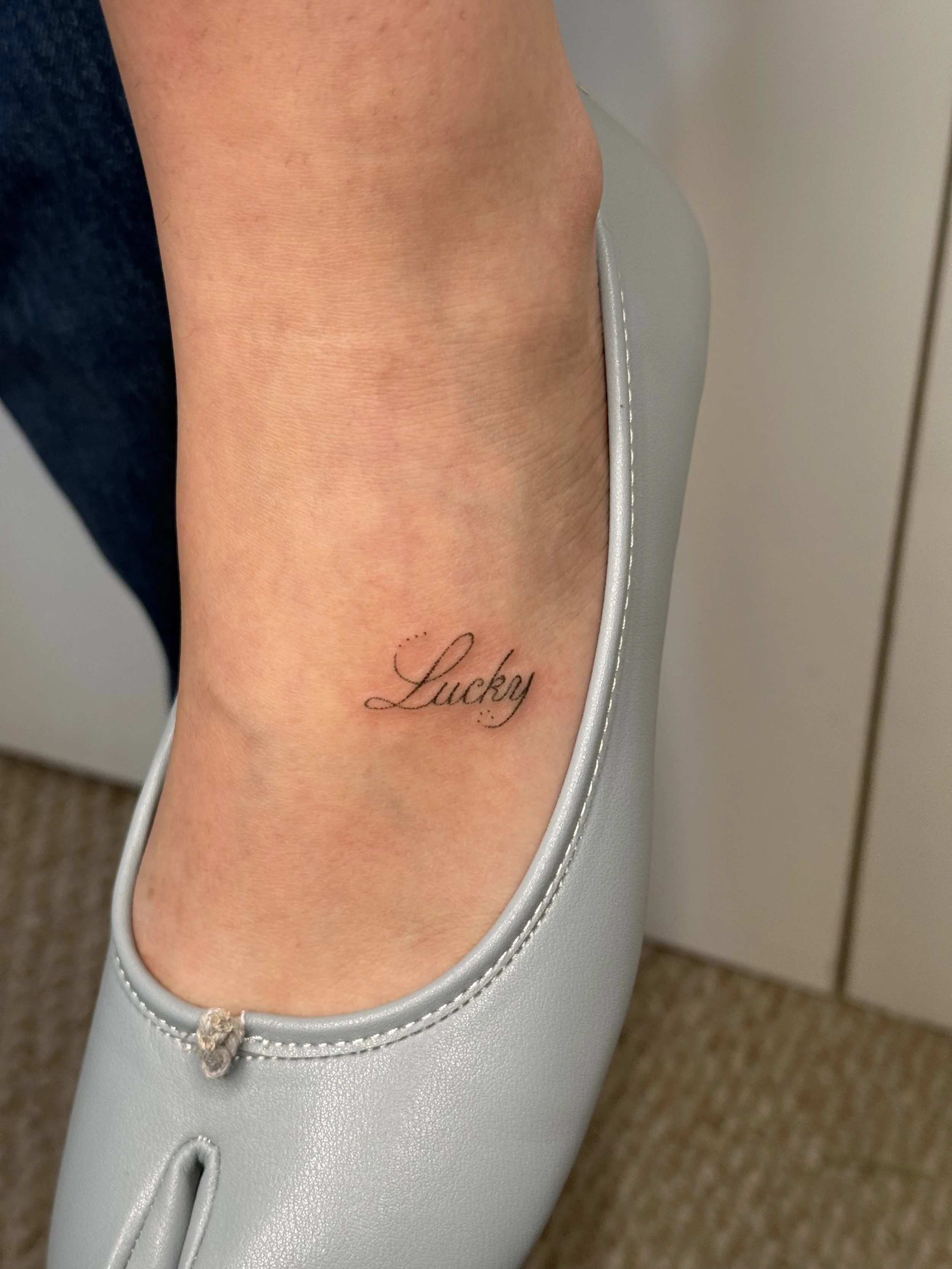 A person's ankle tattoo with the word 'Lucky' written in black cursive, partially covered by a light gray shoe and set against a background of a beige floor and off-white wall.