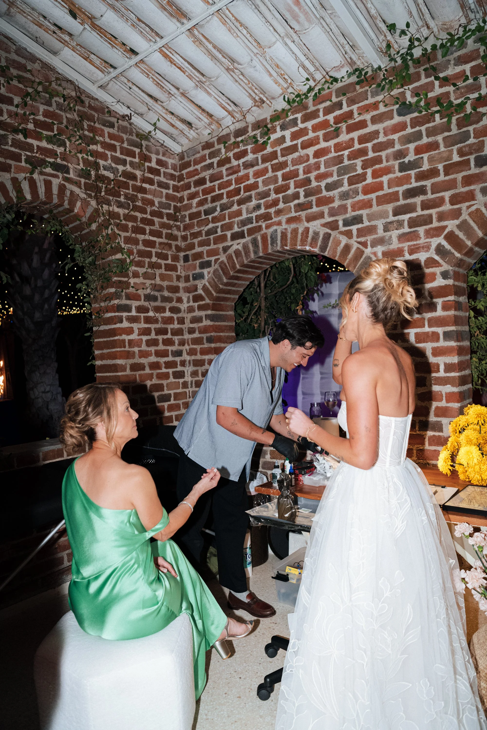 A bride in a white wedding dress standing inside a brick-walled venue, talking to a man in a short-sleeved shirt and black gloves, with women in elegant dresses nearby.