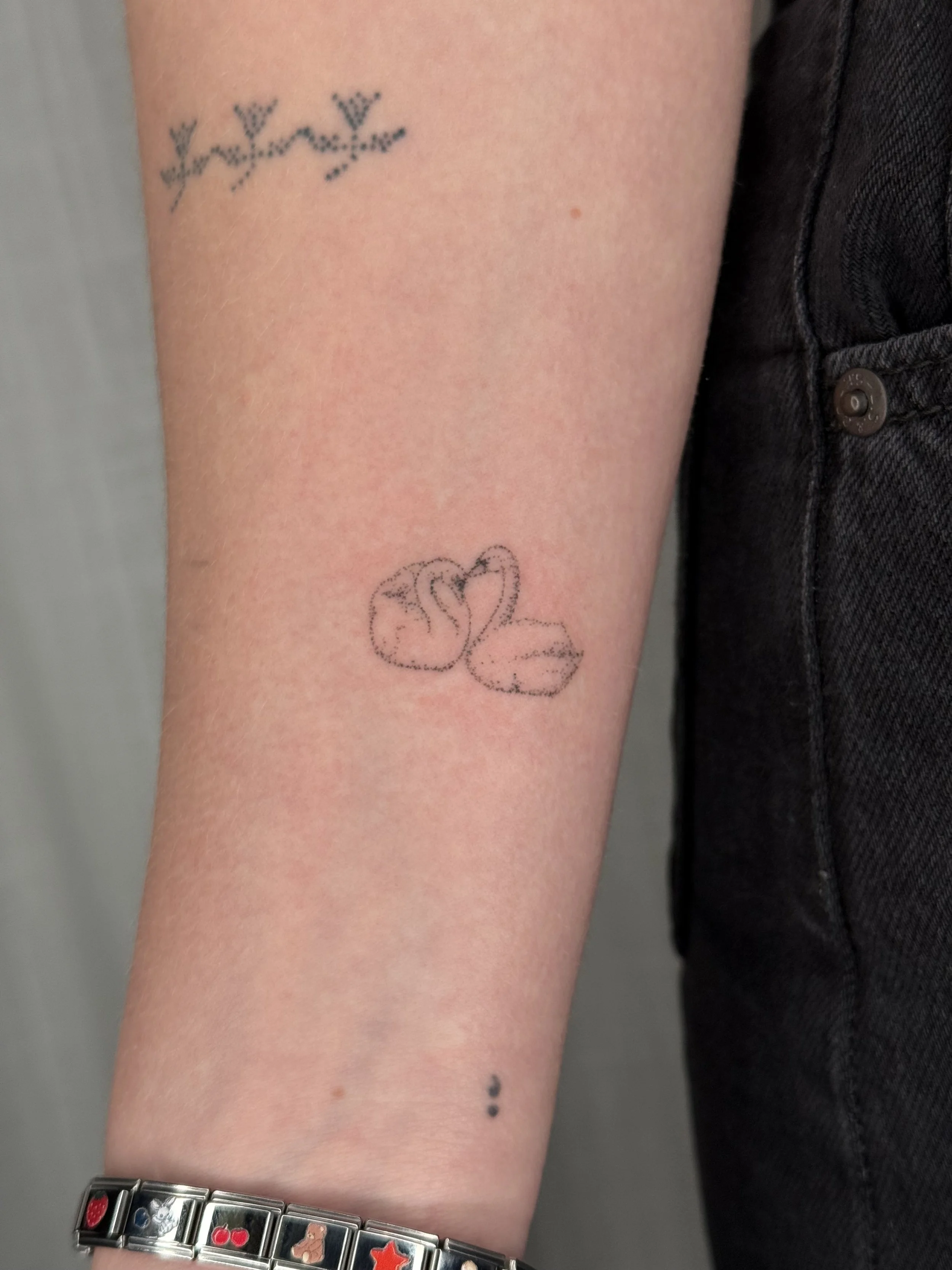 A person's upper arm with three small tattoos, including a heart, stars, and a phrase, and a silver bracelet with colorful charms.