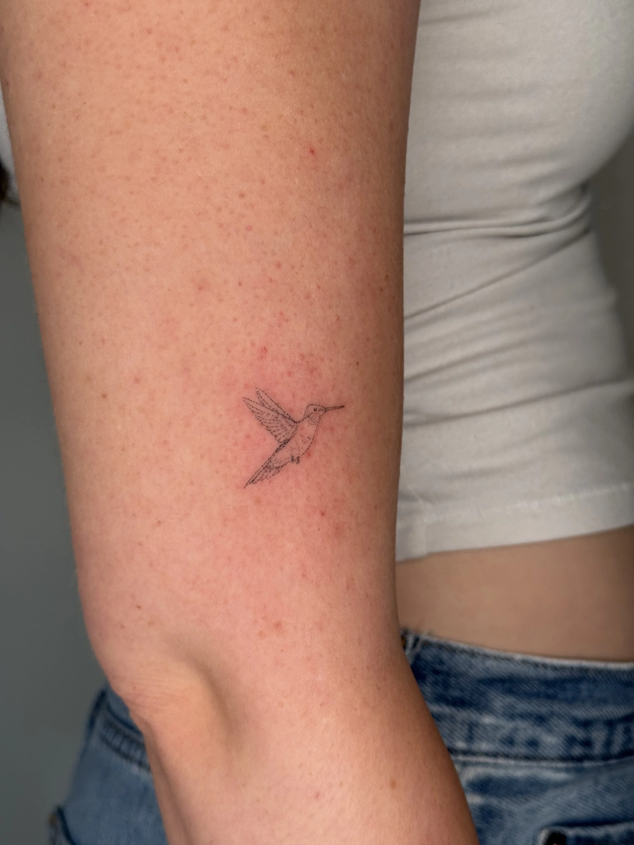 A person's arm with freckles and a small tattoo of a hummingbird near the wrist, wearing a light-colored shirt and sitting next to blue jeans.